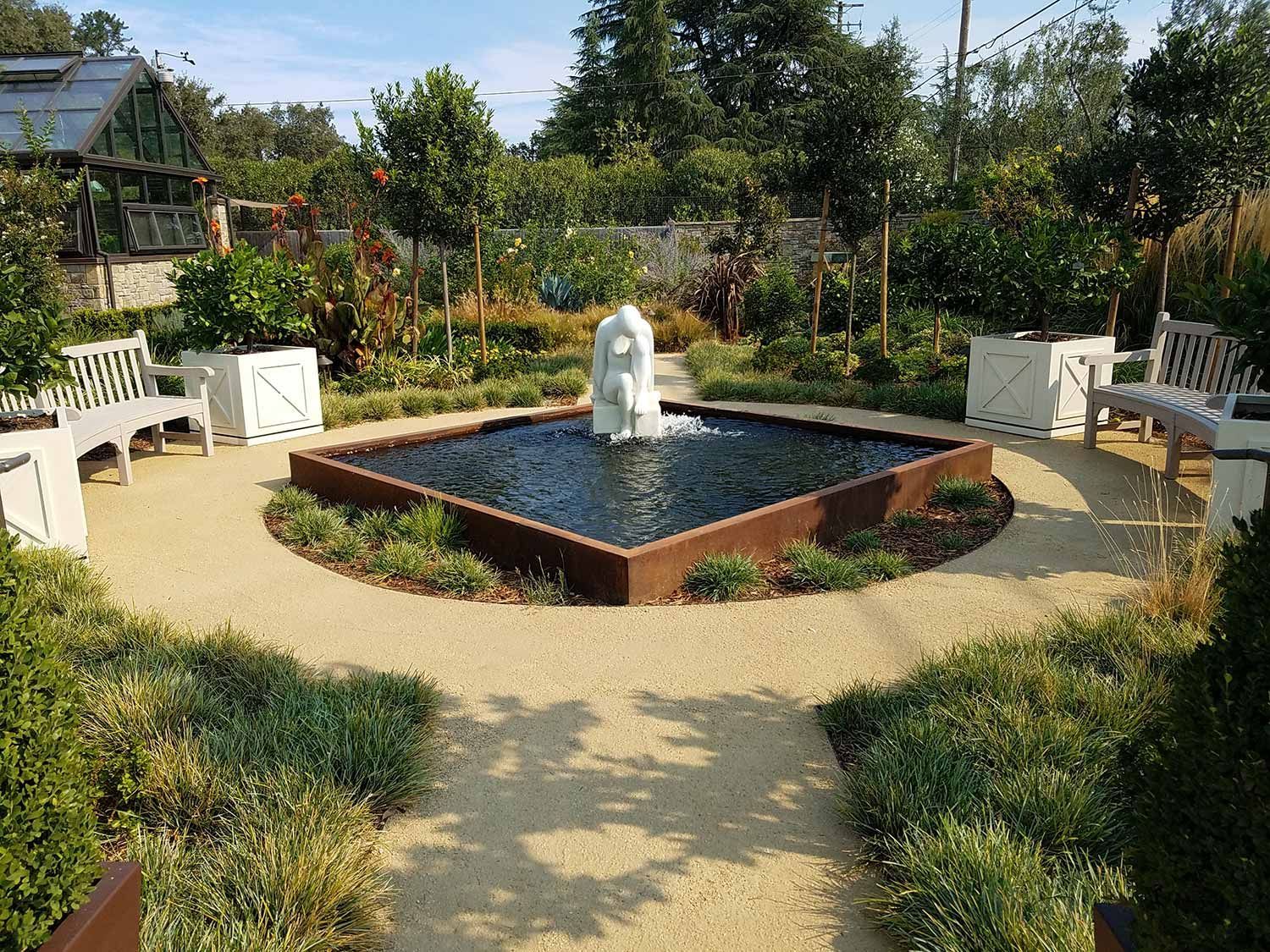 Round garden fountain with white statue, surrounded by benches, flower beds, and a gravel path