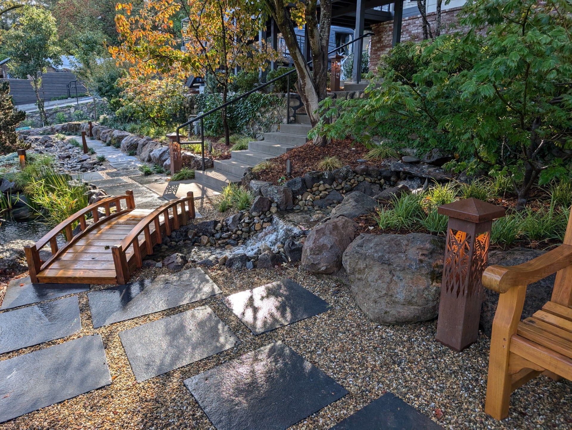 Wooden footbridge over a rocky garden stream beside a gravel path and lush greenery.