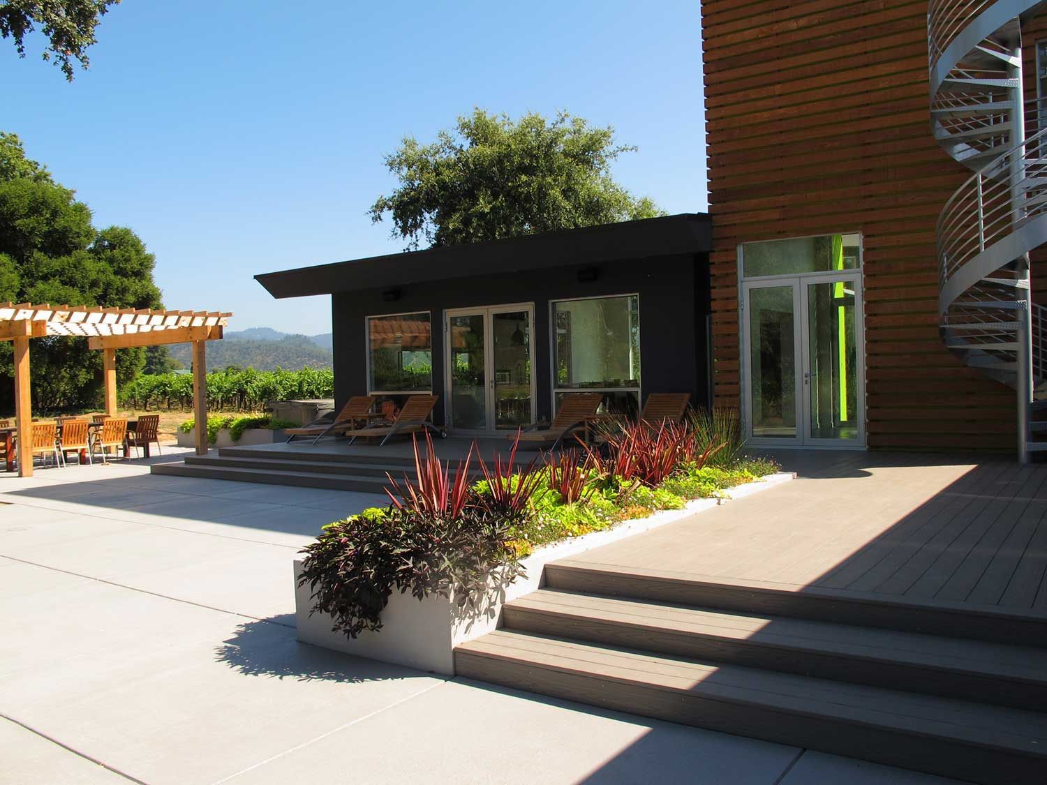 Modern house patio with steps, glass doors, planters, and a spiral staircase beside a sunny courtyard