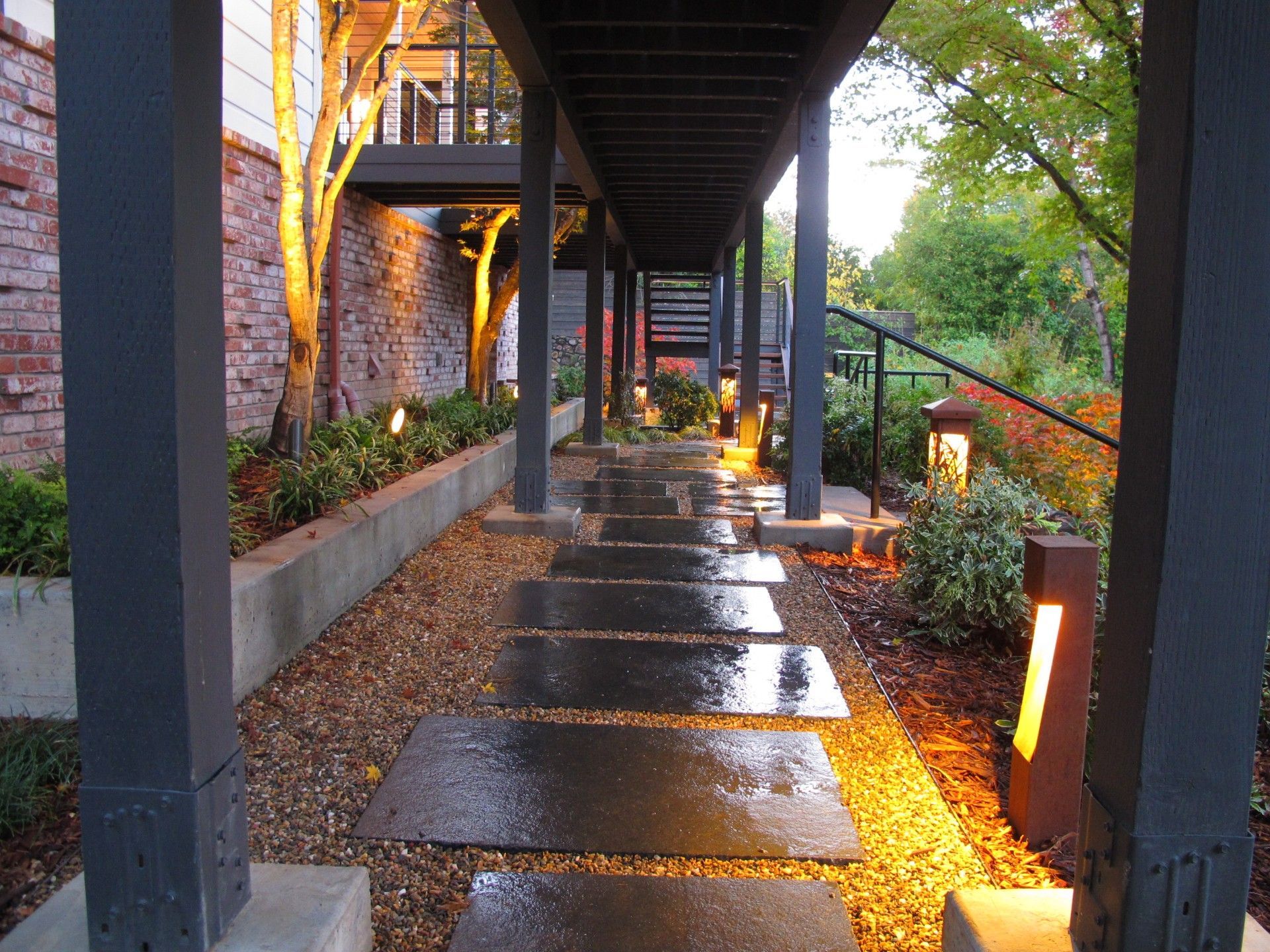 Rainy outdoor walkway with wet stone pavers, covered by a dark awning and lined with warm garden lights.