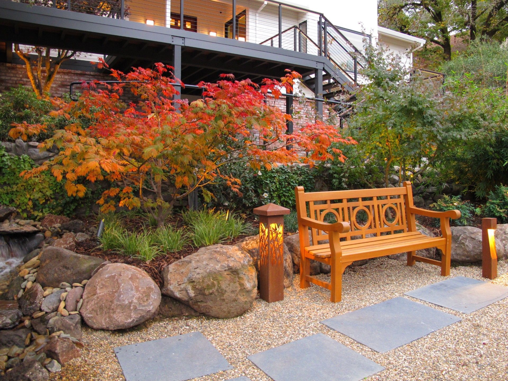 Autumn garden with orange-red shrubs, stone path, and wooden bench beside a deck