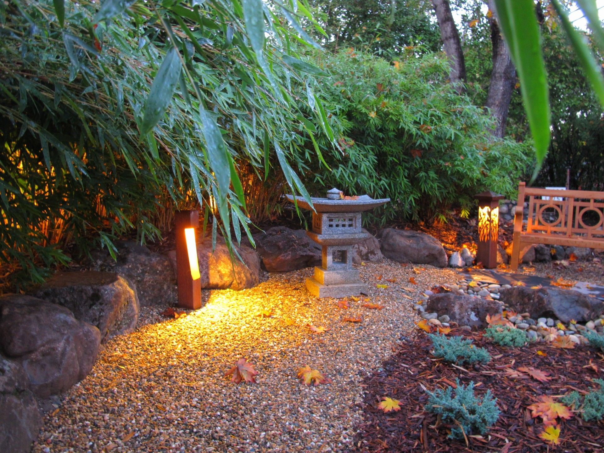 Garden path at dusk with glowing lanterns, gravel ground, dense bamboo, and a stone lantern centerpiece