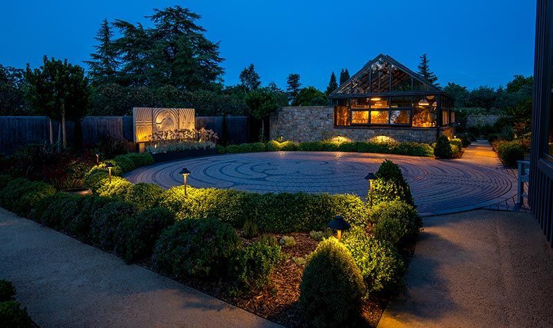 Lit landscaped garden with circular lawn, hedges, and a warmly lit house at dusk