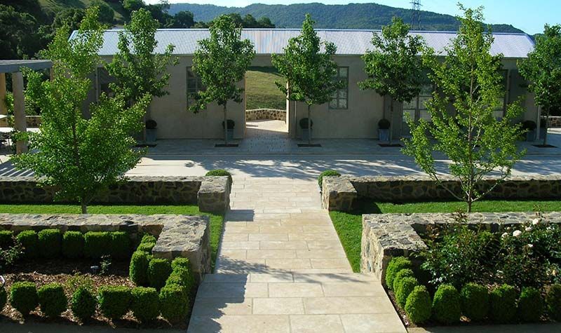 Formal garden path leading to a modern building with trimmed hedges and trees, mountains in the background