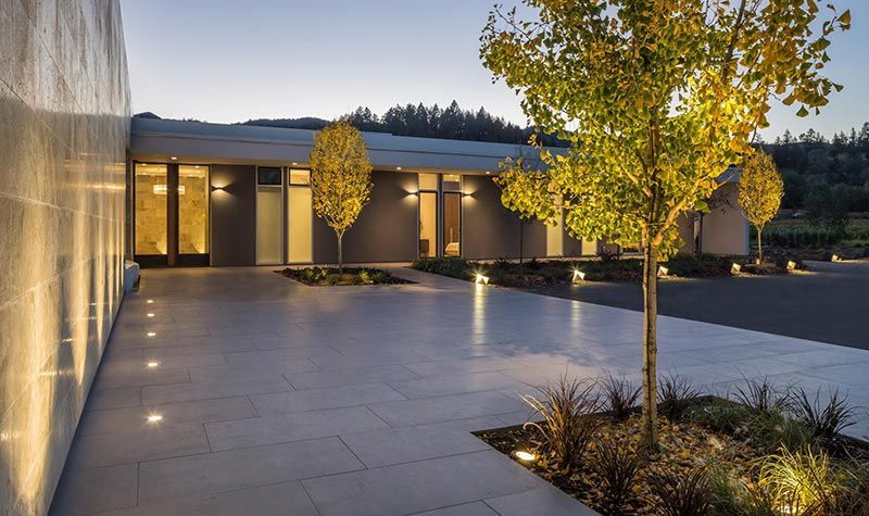Modern courtyard with tiled paving, lit glass walls, and yellow trees at dusk