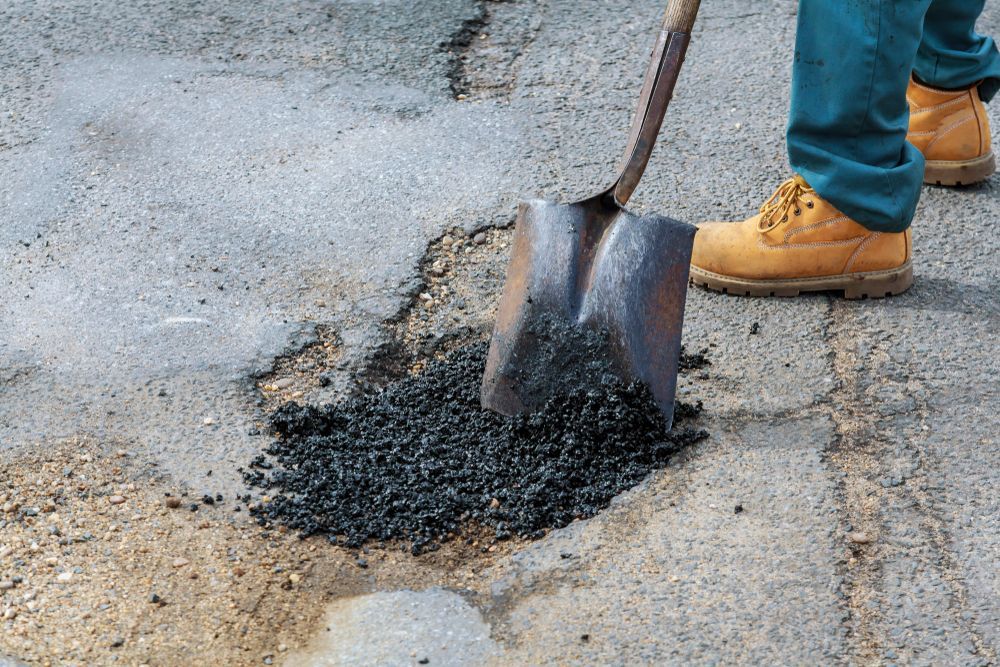 Shovel spreading black asphalt patch on a damaged road beside a worker in boots