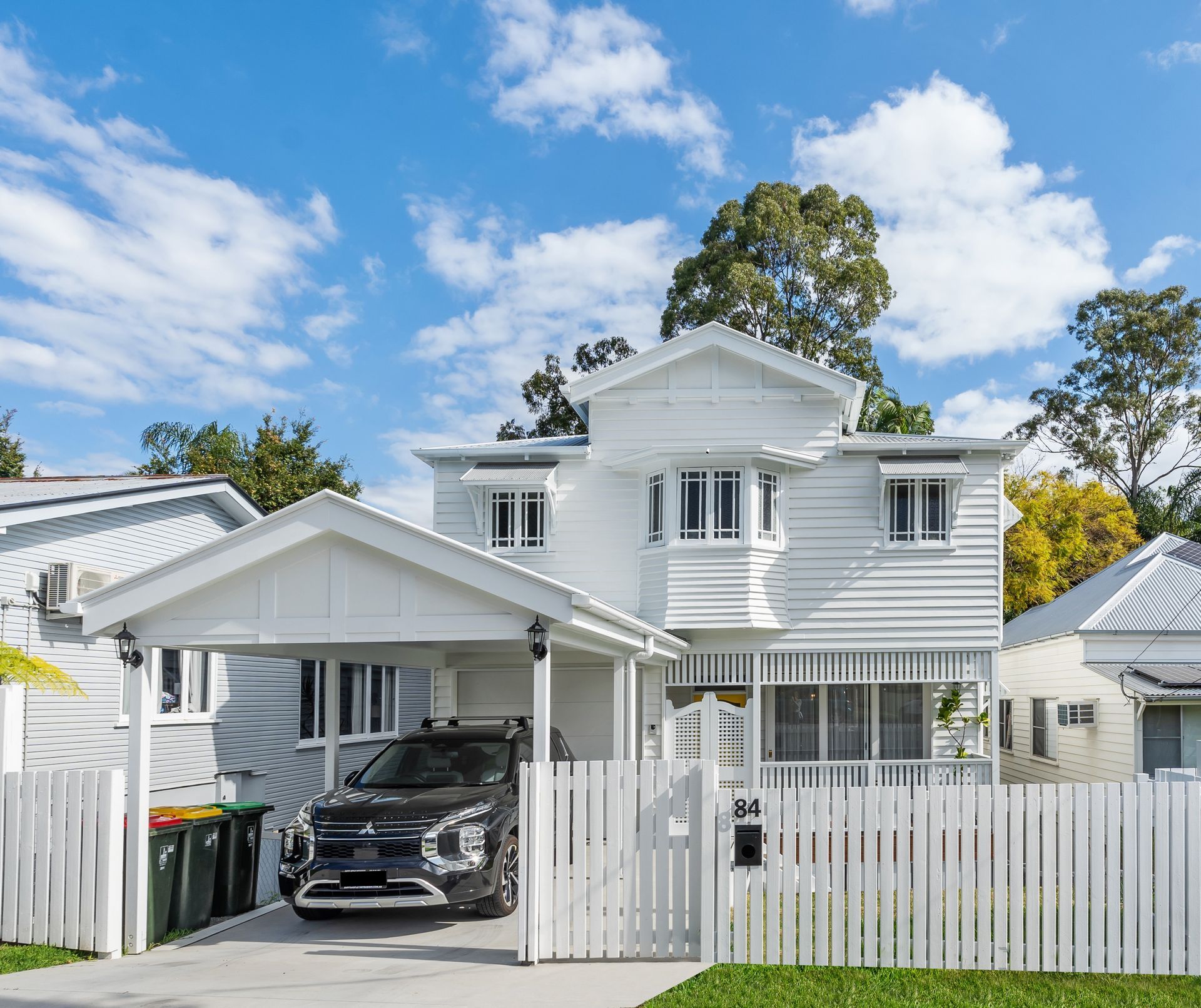 White two-story house with a carport, black SUV, and white picket fence under a blue sky