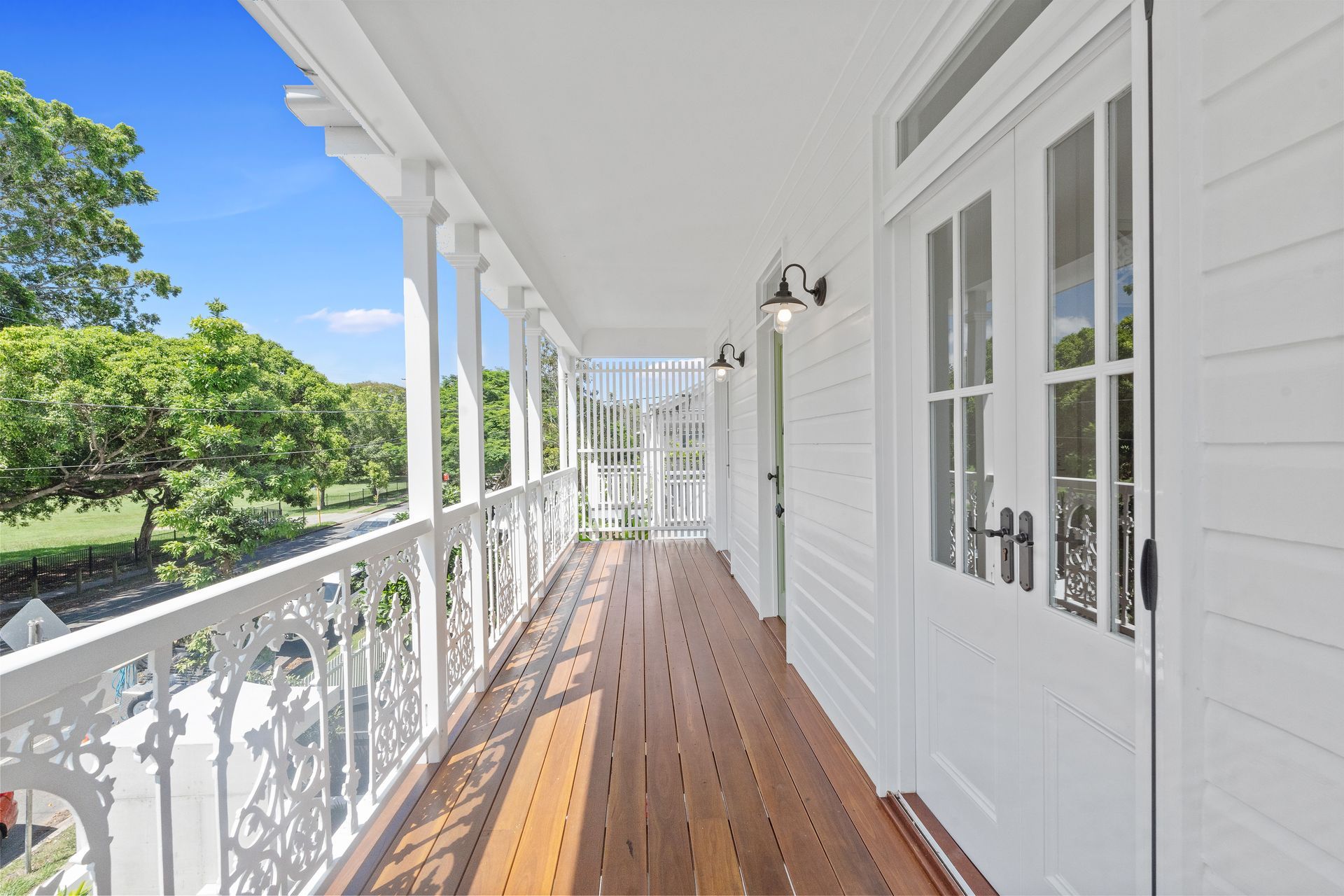 White porch with wooden floor, ornate railing, and French doors overlooking green trees on a sunny day