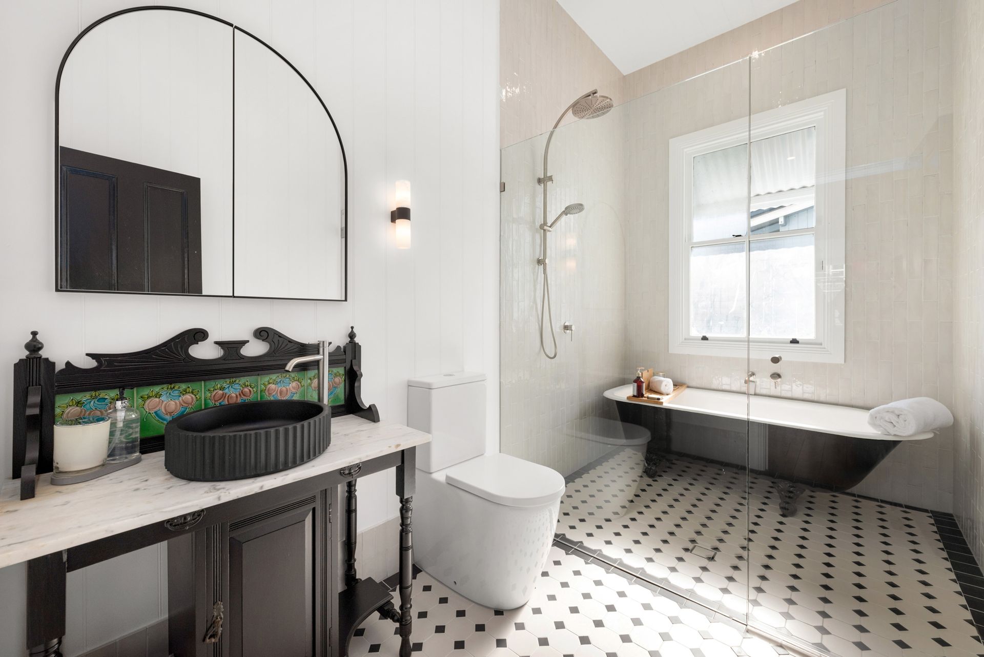 Modern bathroom with black-and-white tiled floor, freestanding tub, toilet, vanity, and arched mirror.