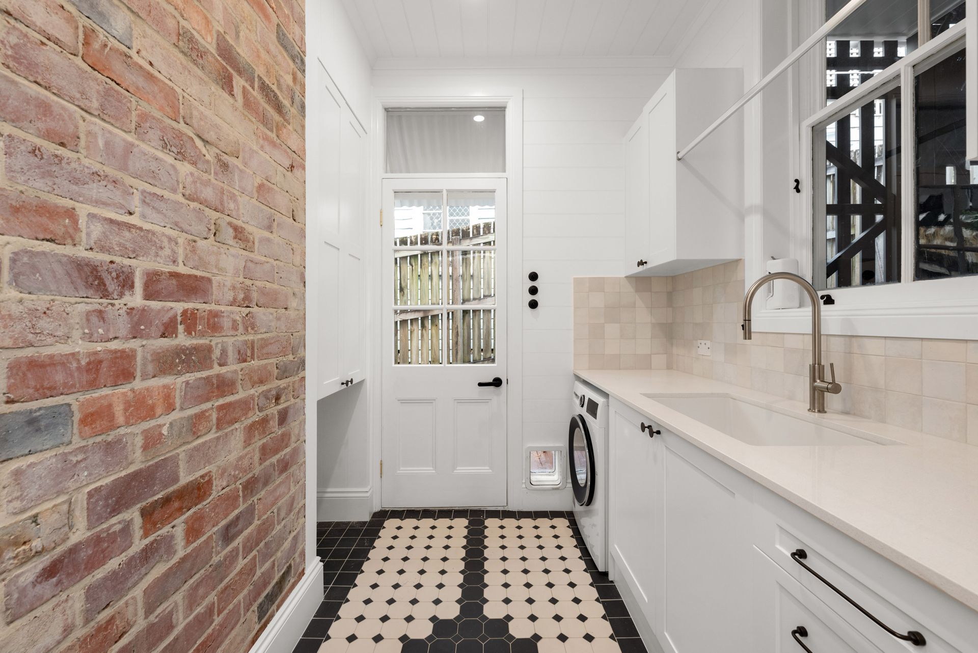 Narrow white kitchen with brick wall, black-and-white tile floor, and a glass-paneled door at the end.