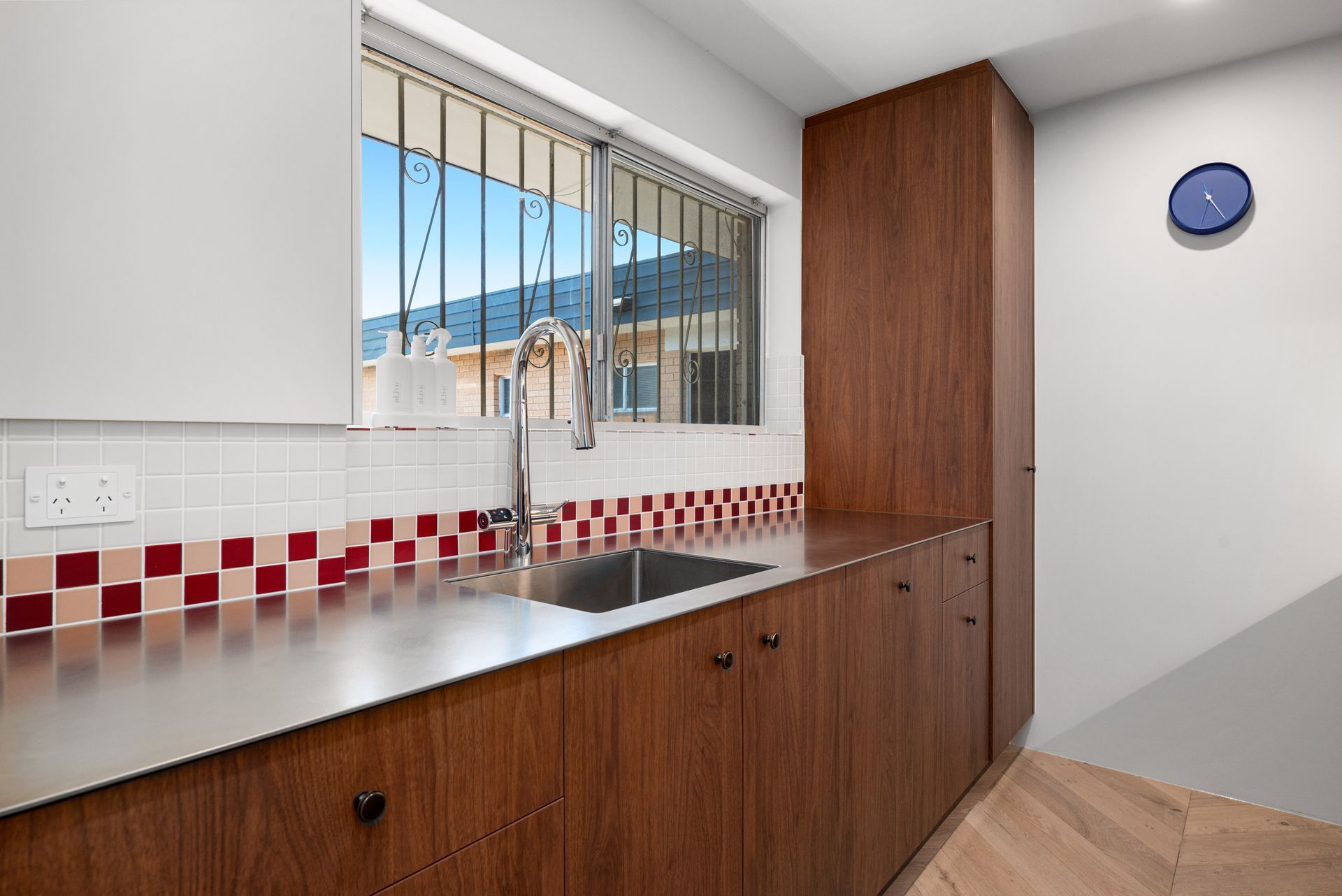 Modern kitchen with wooden cabinets, stainless countertop, red-and-white tile backsplash, sink, and window with bars