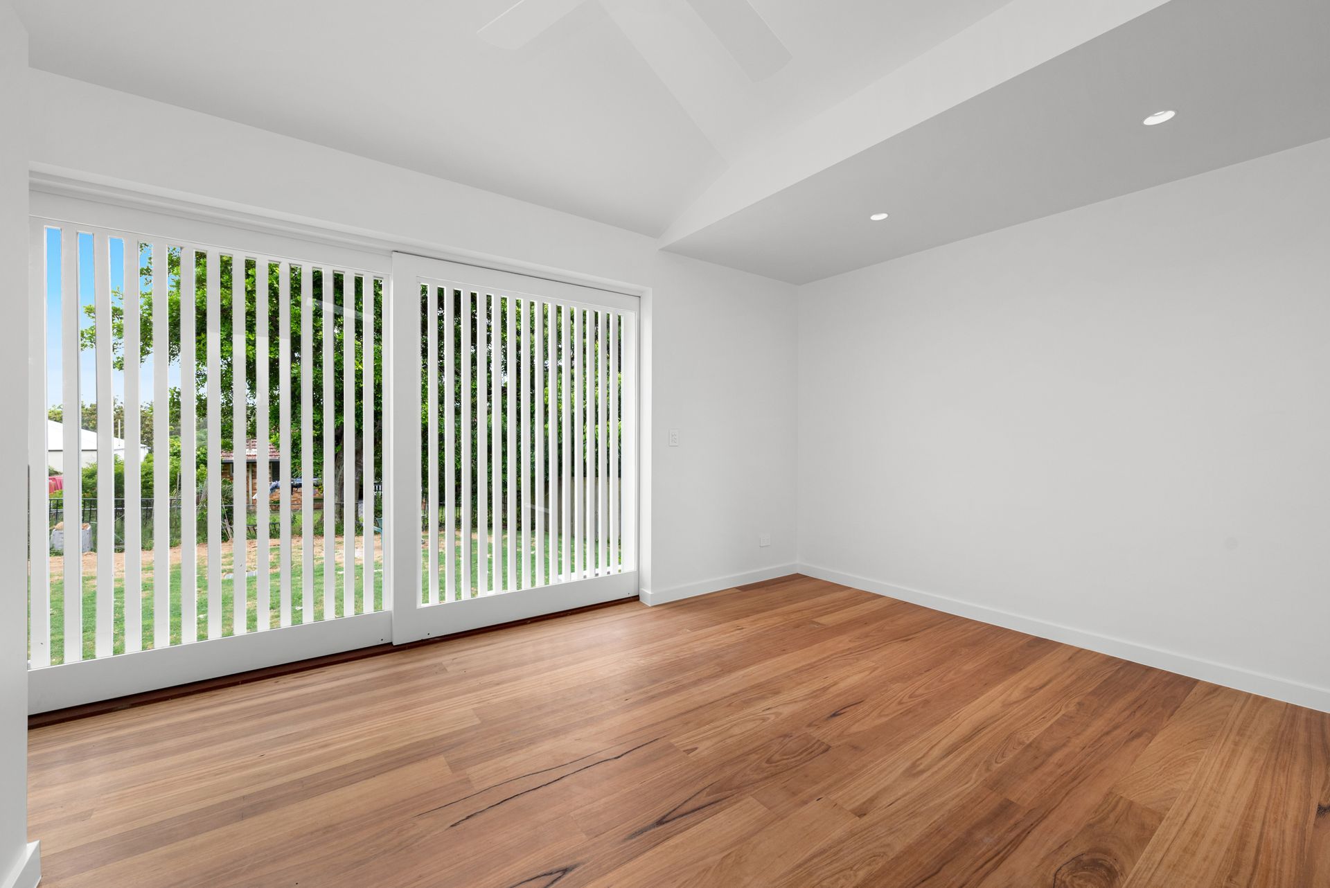 Empty bright room with hardwood floors, white walls, and large vertical blinds letting in daylight.