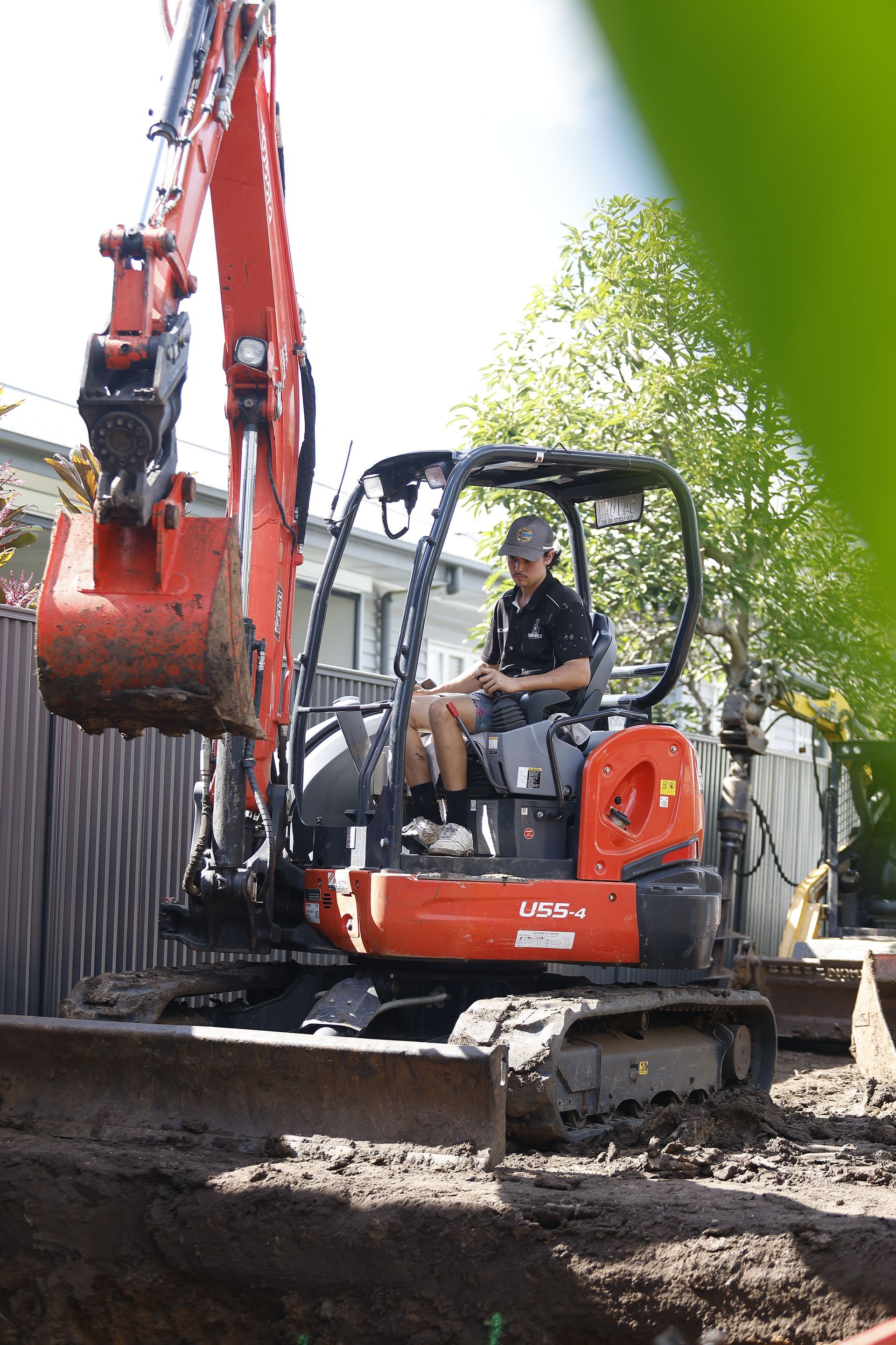 Excavator digging in a dirt trench beside a fence, with a worker operating it near trees.