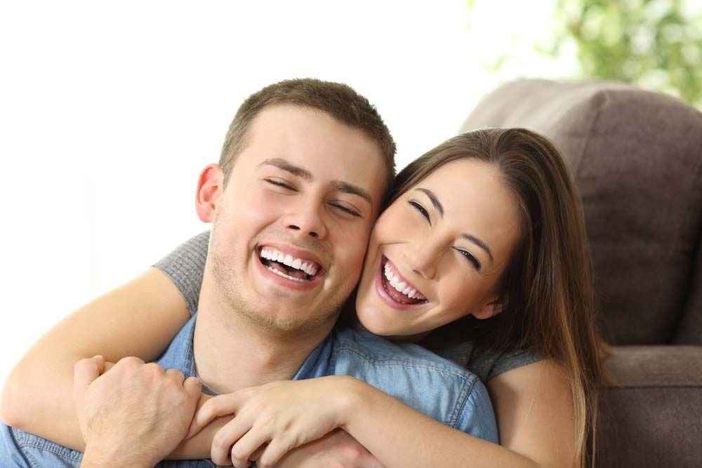 A Man and A Woman Are Hugging Each Other and Smiling While Sitting on A Couch — Teeth Whitening in Newcastle, NSW