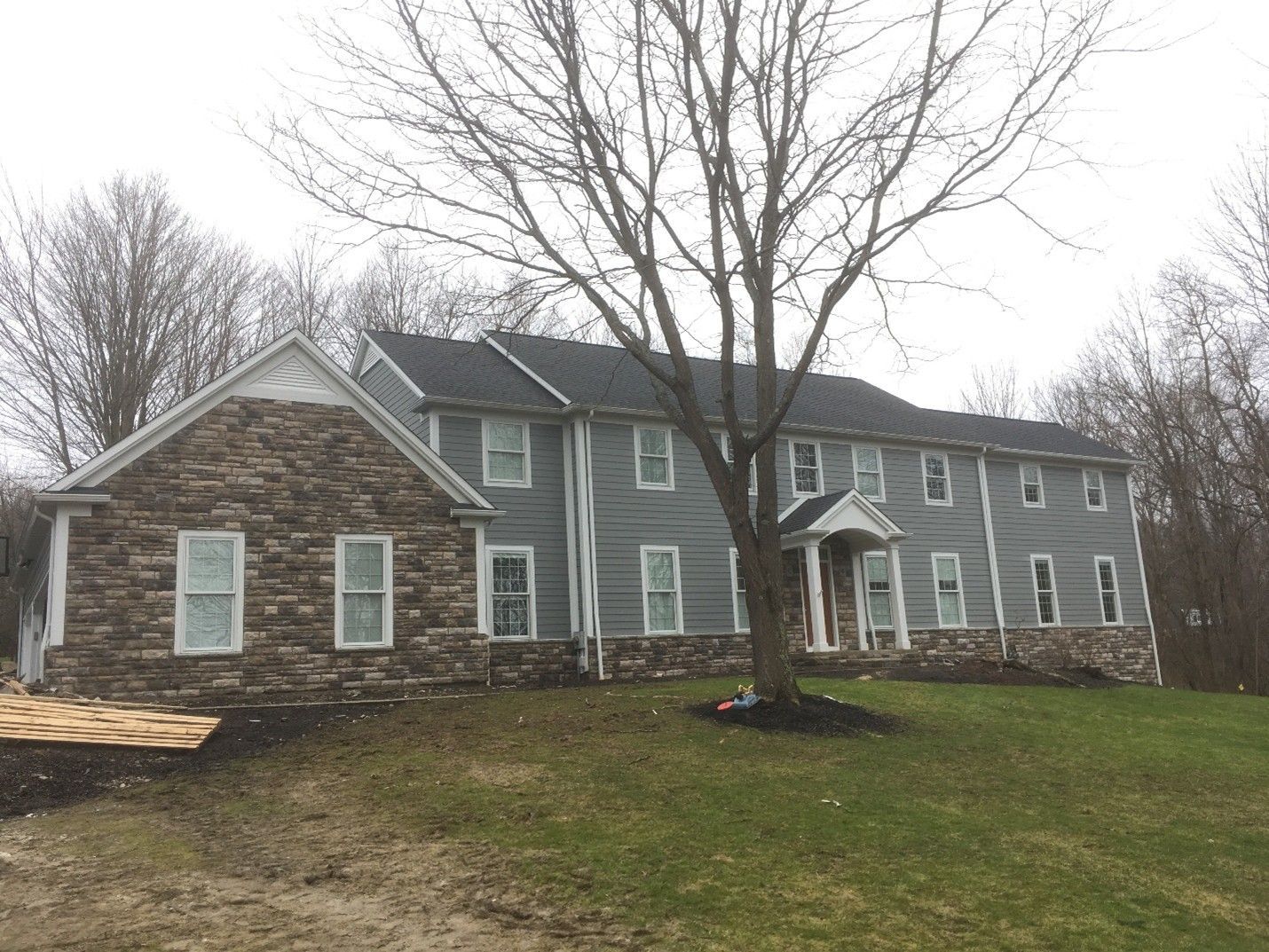 Two-story gray house with stone facade, dark roof, windows, and a tree in front on a green hill.