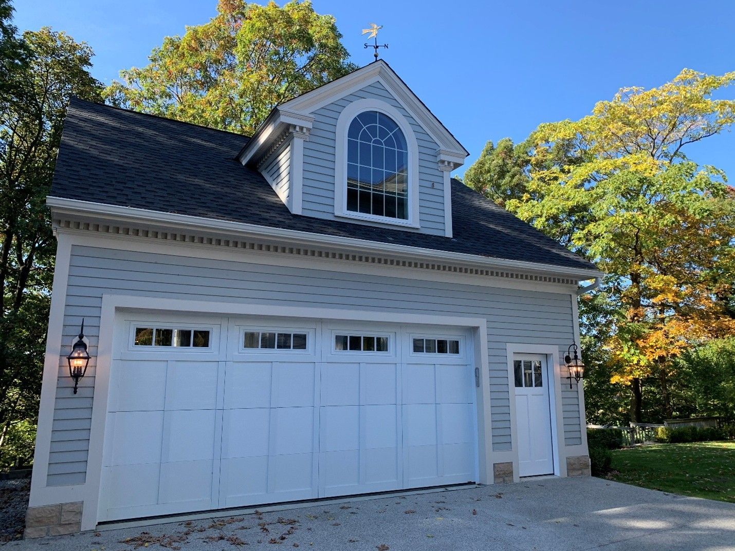 Light blue two-story garage with white doors, arched window, and weathervane against a blue sky.