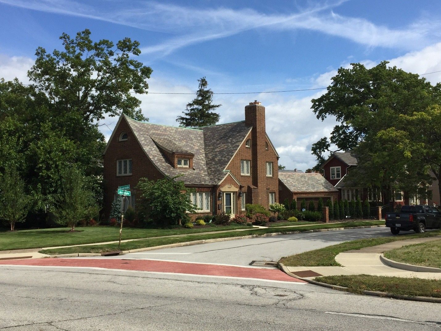 Brick house with a steeply pitched roof, chimney, and landscaped lawn on a corner lot under a blue sky.