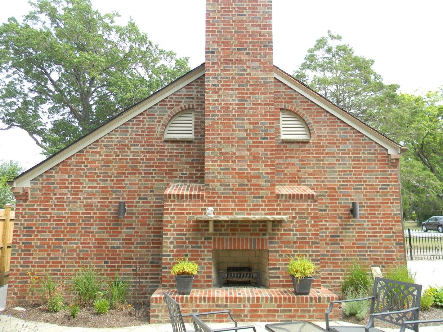 Brick outdoor fireplace with chimney. Brown and green plants in pots and surrounding the base of the fireplace.
