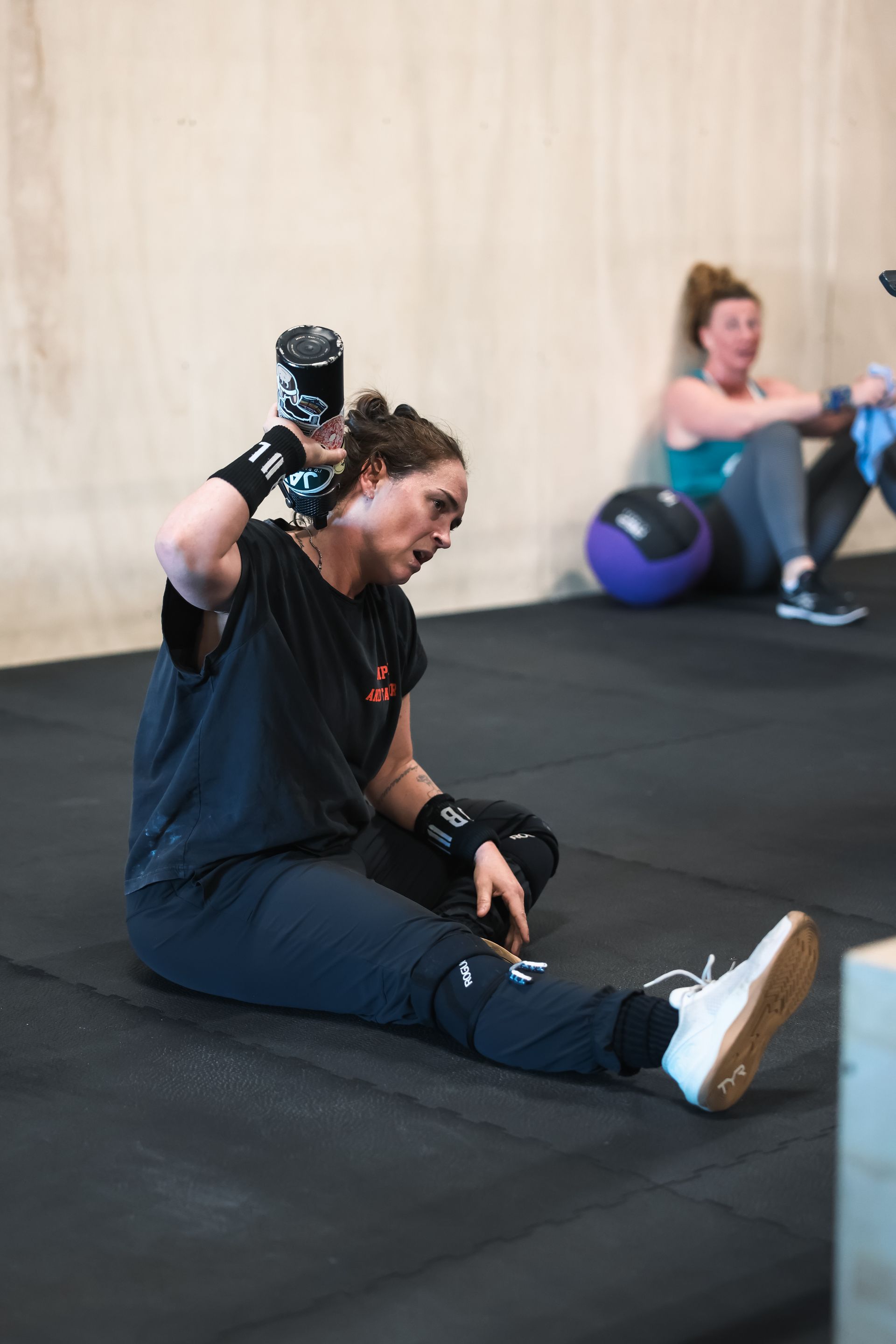 Woman stretching on gym floor while holding a dumbbell overhead, with another person exercising in the background