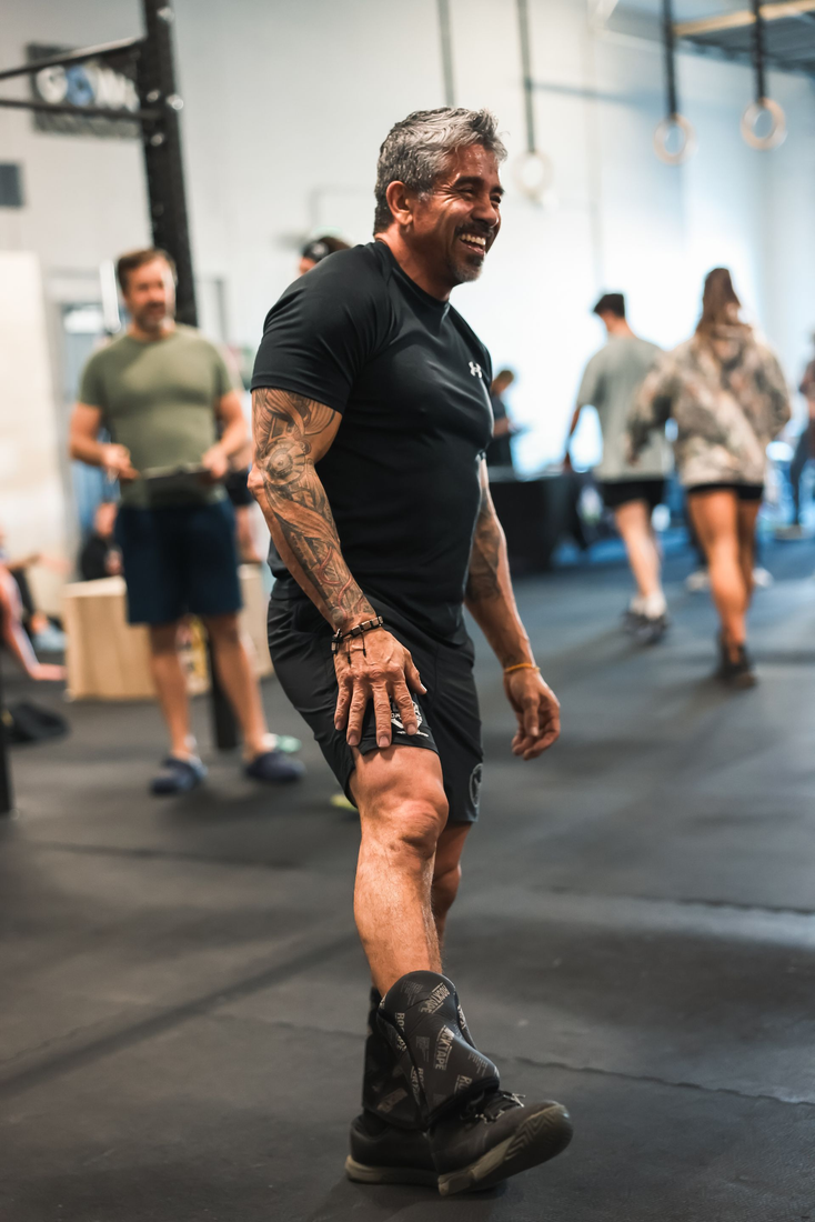 Man in black workout clothes standing in a gym, with other people exercising in the background.