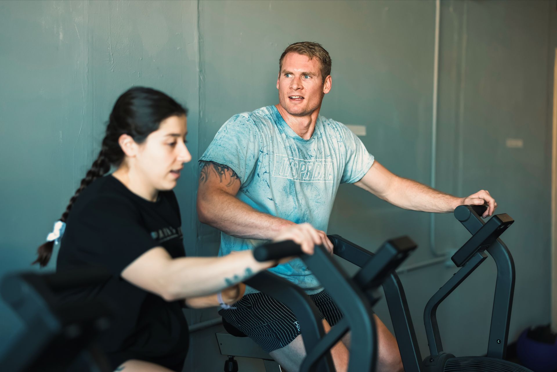 Two people exercising on stationary bikes in a gym in Northglenn, CO, one focused and one talking while riding.
