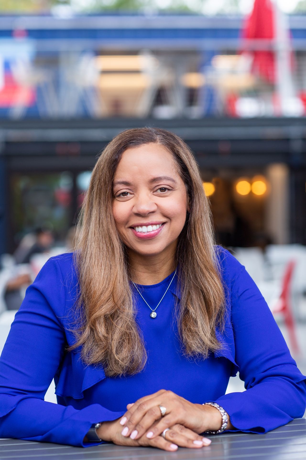 A woman in a blue shirt is sitting at a table with her hands folded and smiling.