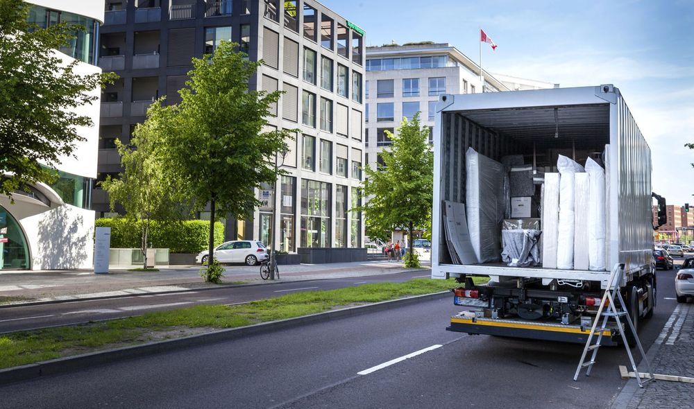 Un camion scarica pannelli bianchi alti su una strada cittadina, accanto a edifici moderni e alberi.