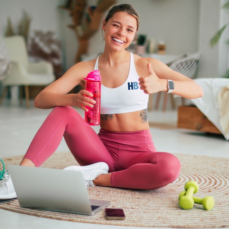 A woman is sitting on the floor holding a water bottle and giving a thumbs up. Achieve Results with HomeBodies HIIT Personal Training