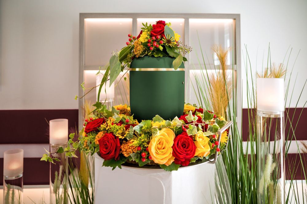 A green funeral urn rests on a white pedestal, surrounded by a floral wreath — Ashes to Ashes Cremations In Port Douglas, QLD