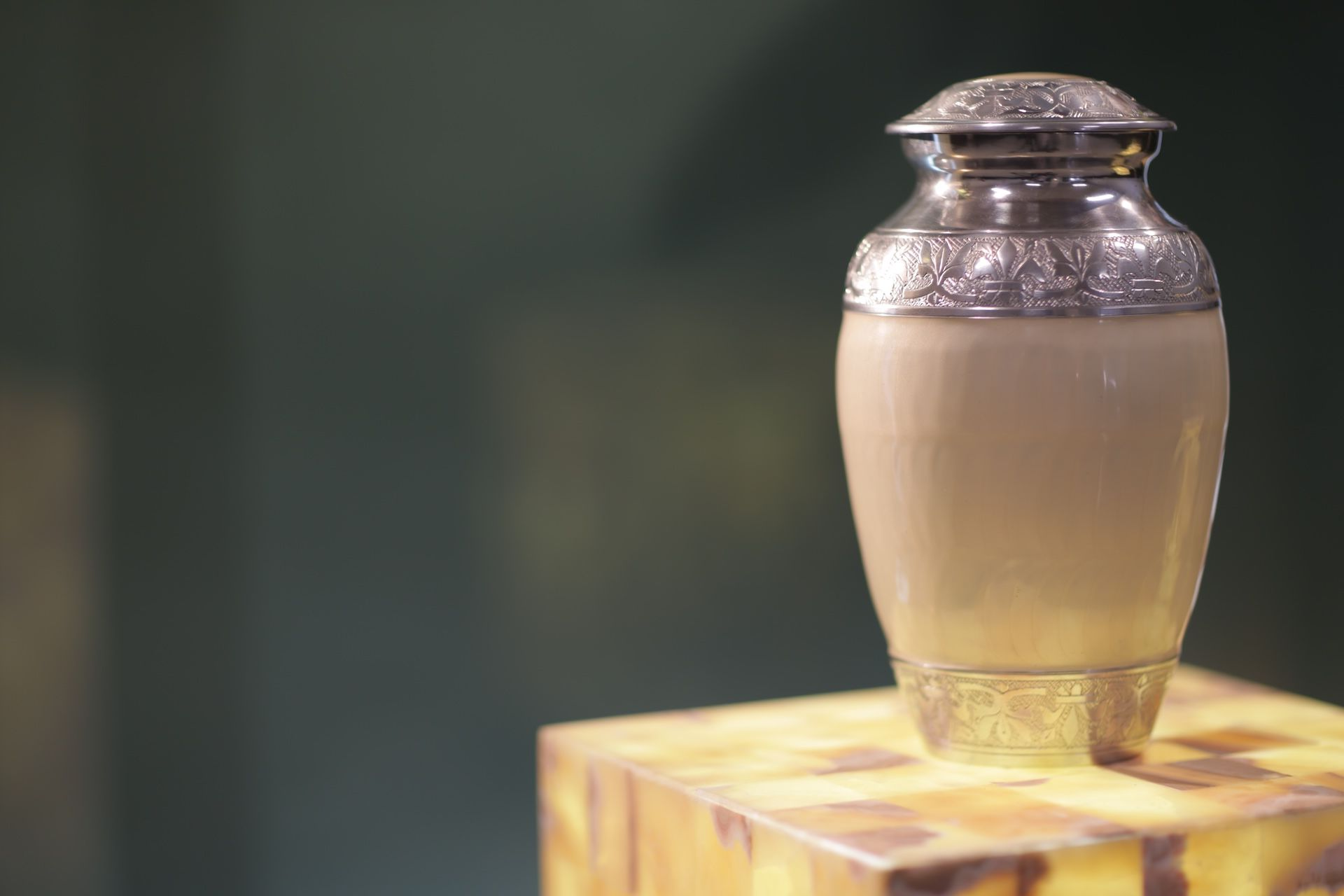 A silver urn-shaped vase with a cream base sits on a wooden pedestal against a blurred green background — Ashes to Ashes Cremations In Tablelands, QLD