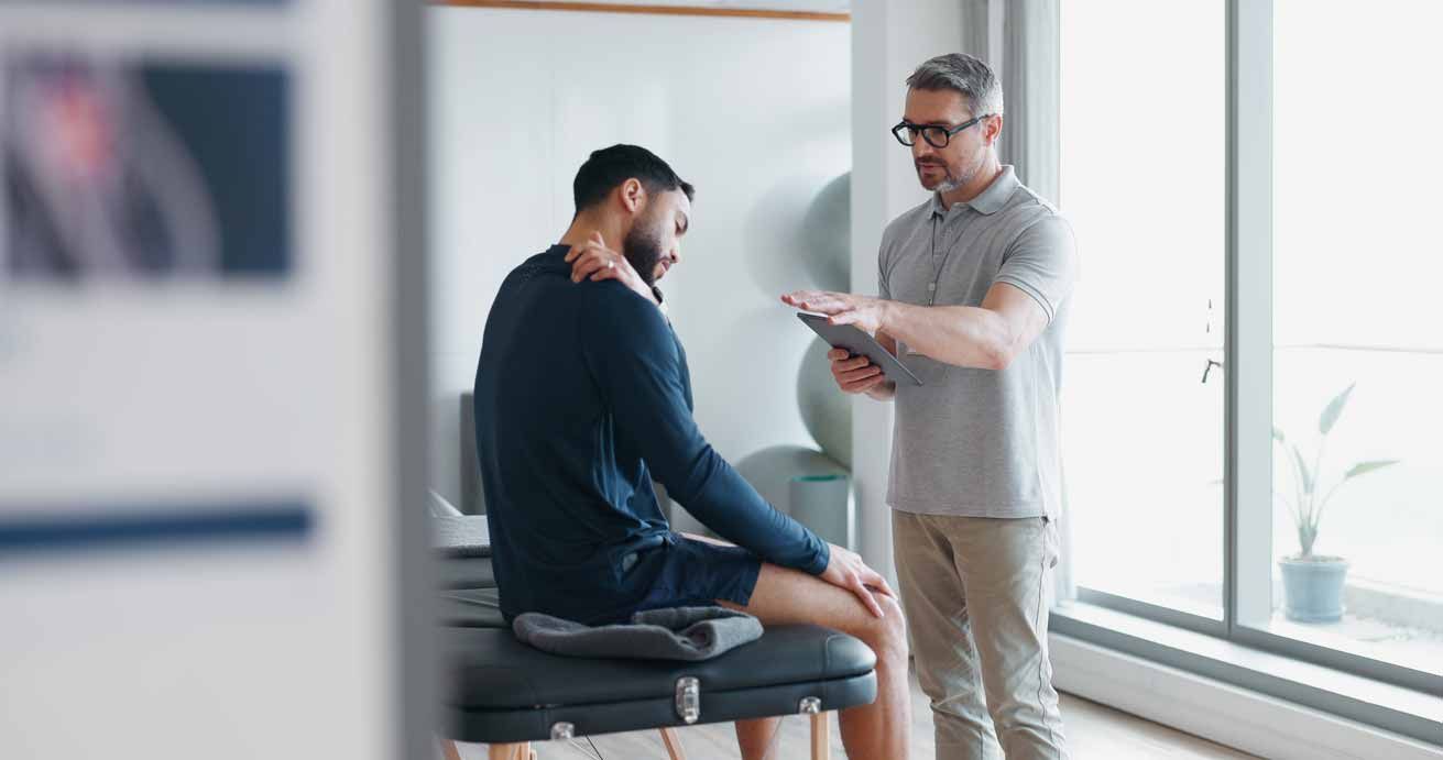 Two men in a bright clinic, one seated on an exam table while the other checks his arm with a paper chart