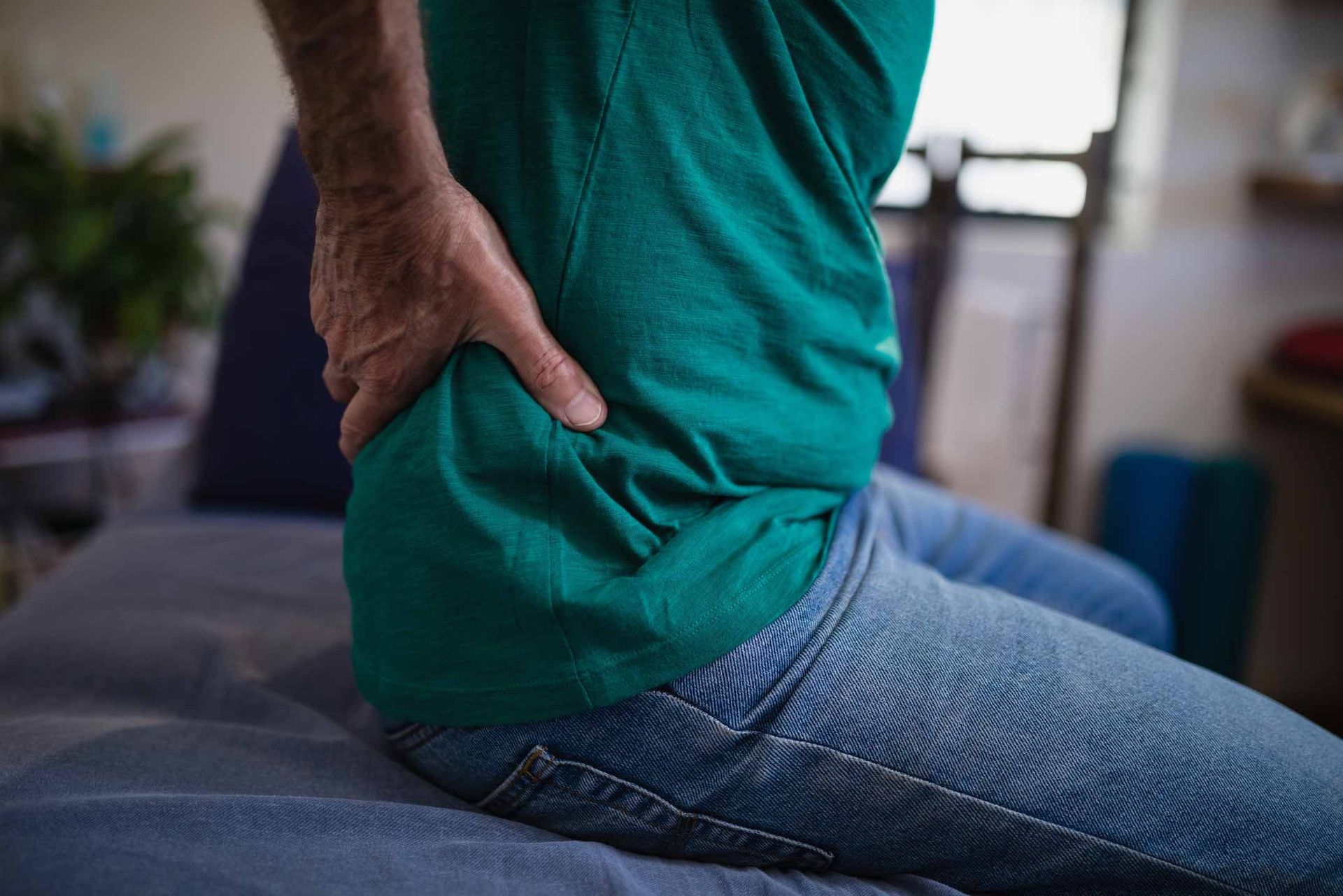 Person seated on a bed, holding their lower back with a hand in a teal shirt and jeans.