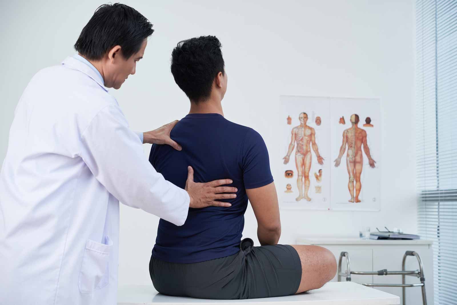 Doctor examining a seated patient’s back in a bright clinic, with anatomical charts on the wall