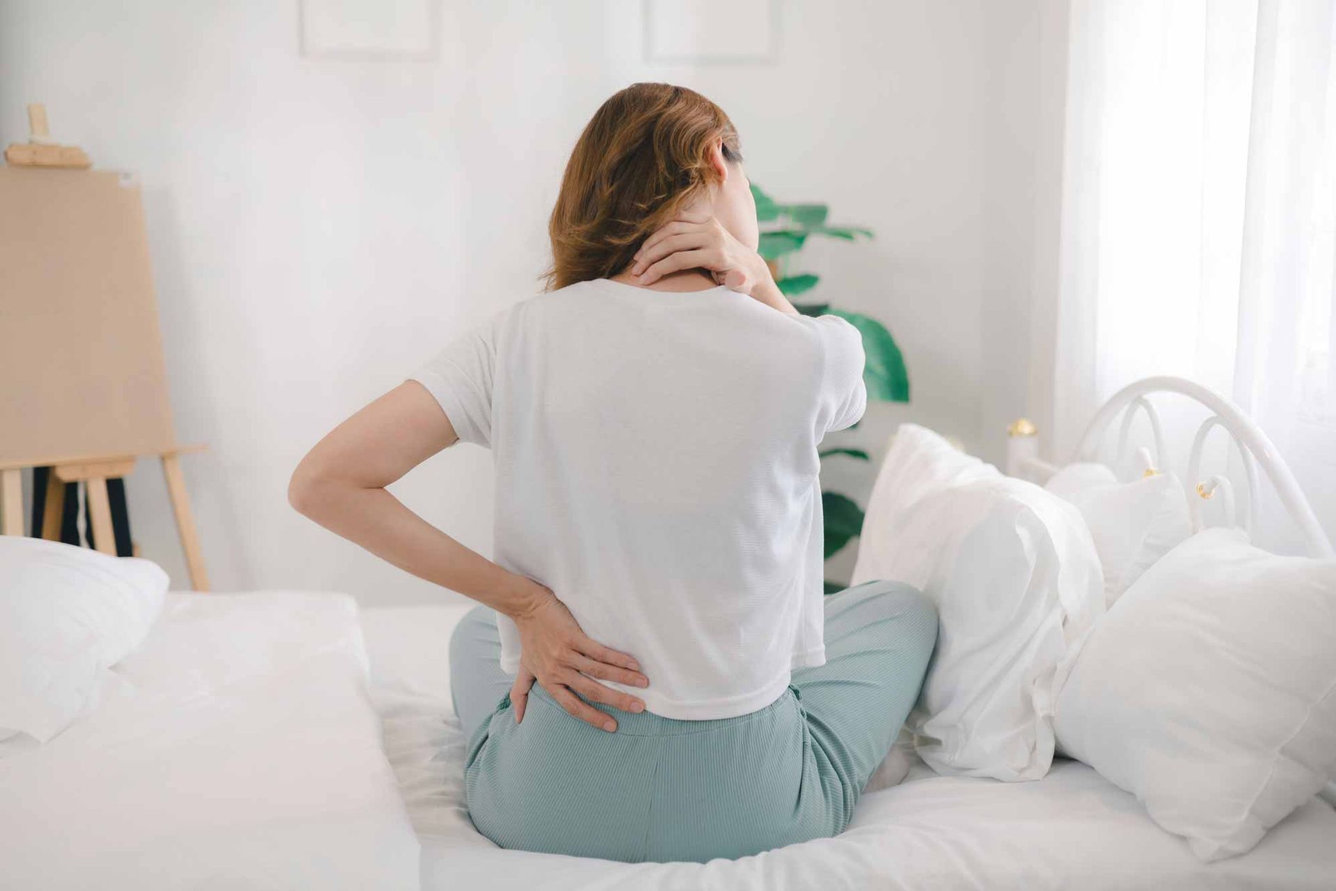 Person sitting on a bed and holding their lower back, suggesting back pain in a bright bedroom.