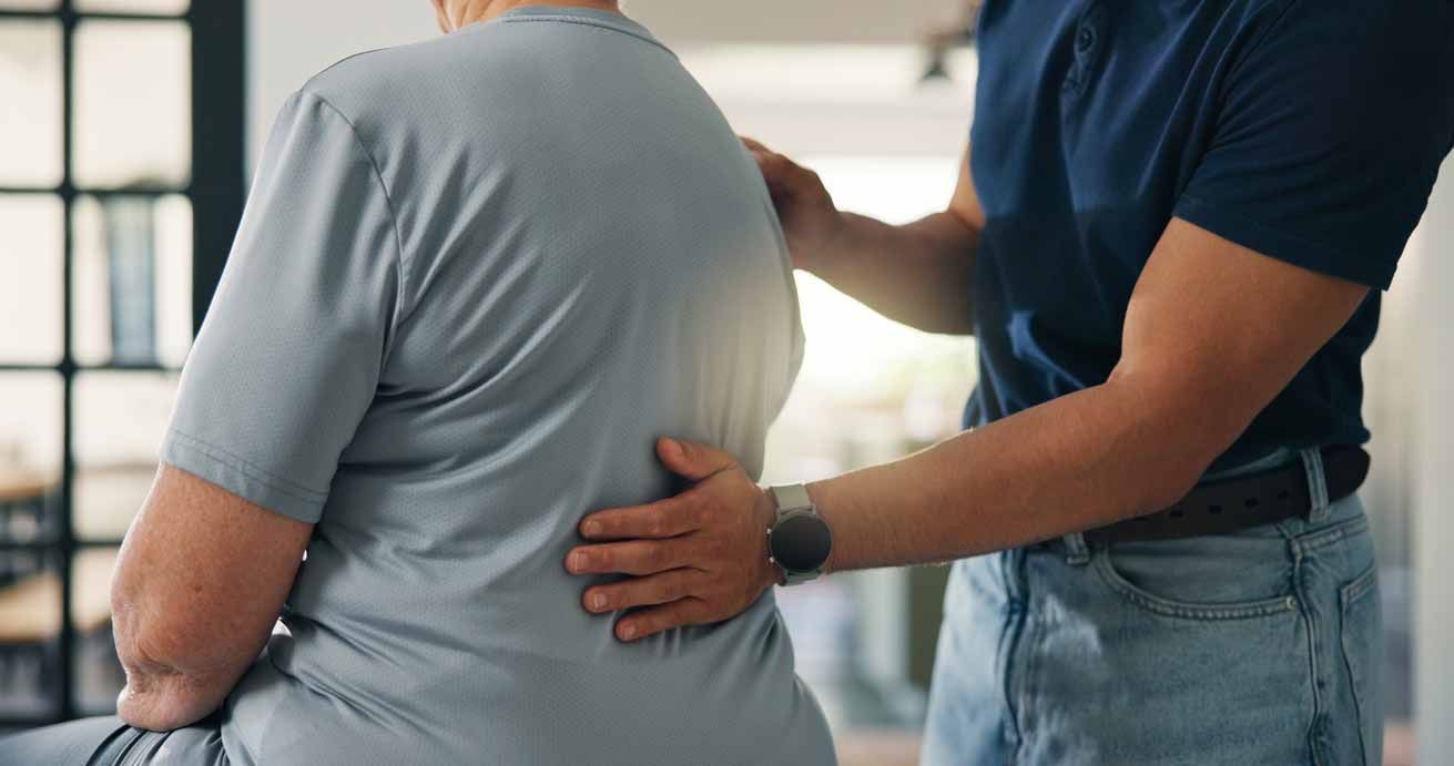 Caregiver places a hand on a seated person’s back in a bright indoor setting.