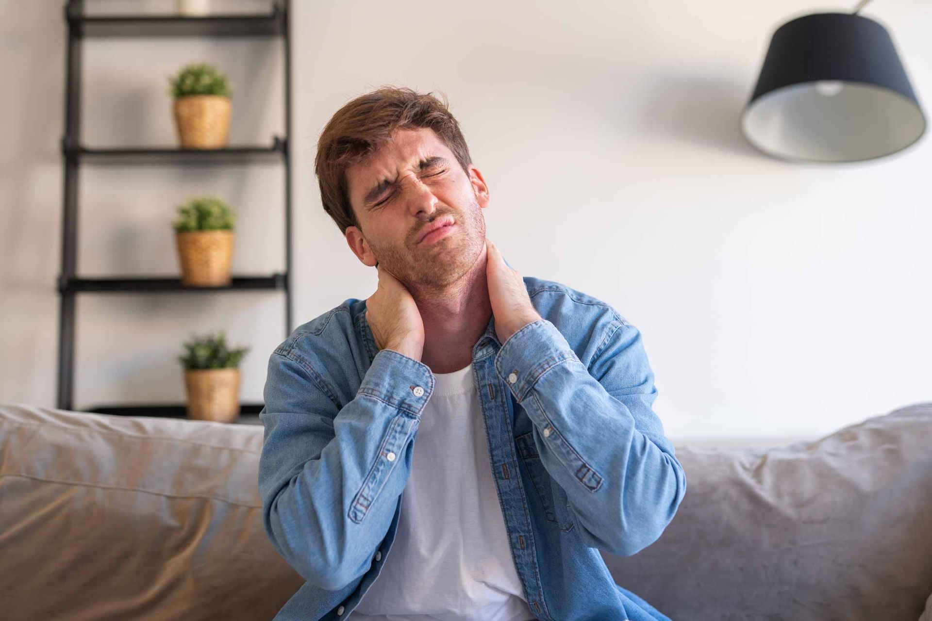 Person sitting on a couch, rubbing their neck and looking uncomfortable in a bright living room.