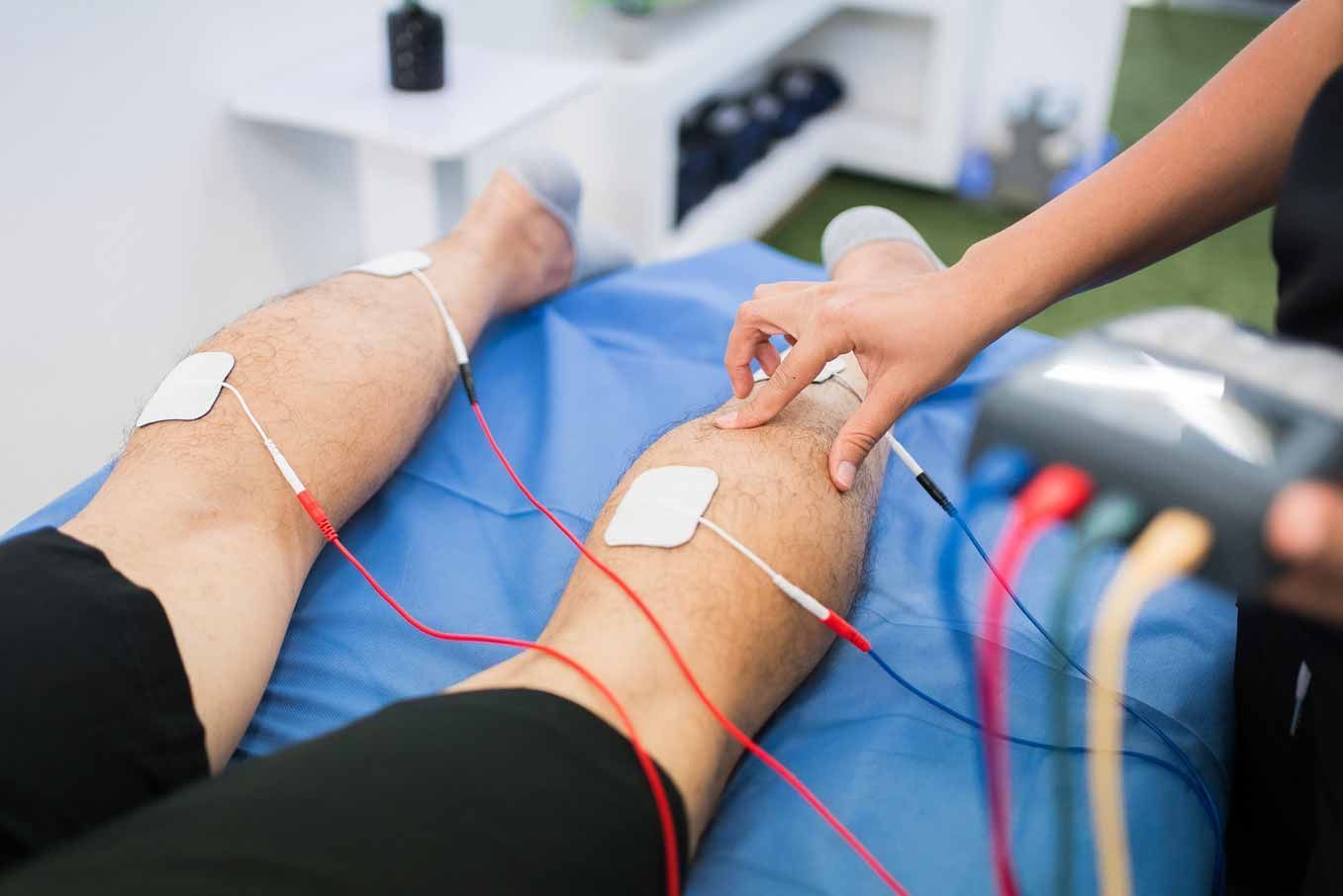 Physical therapy session with electrodes on a patient’s legs and a therapist holding one knee