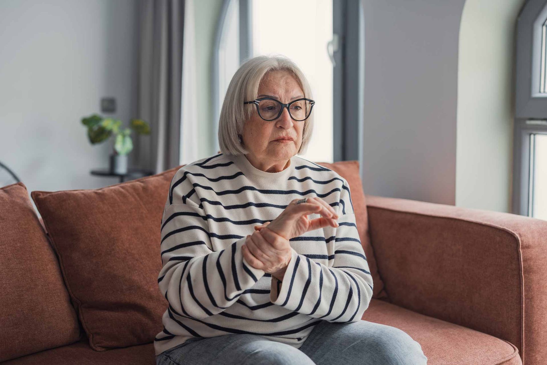 Older woman sitting on a couch, wearing glasses and a striped shirt, looking thoughtful by a window