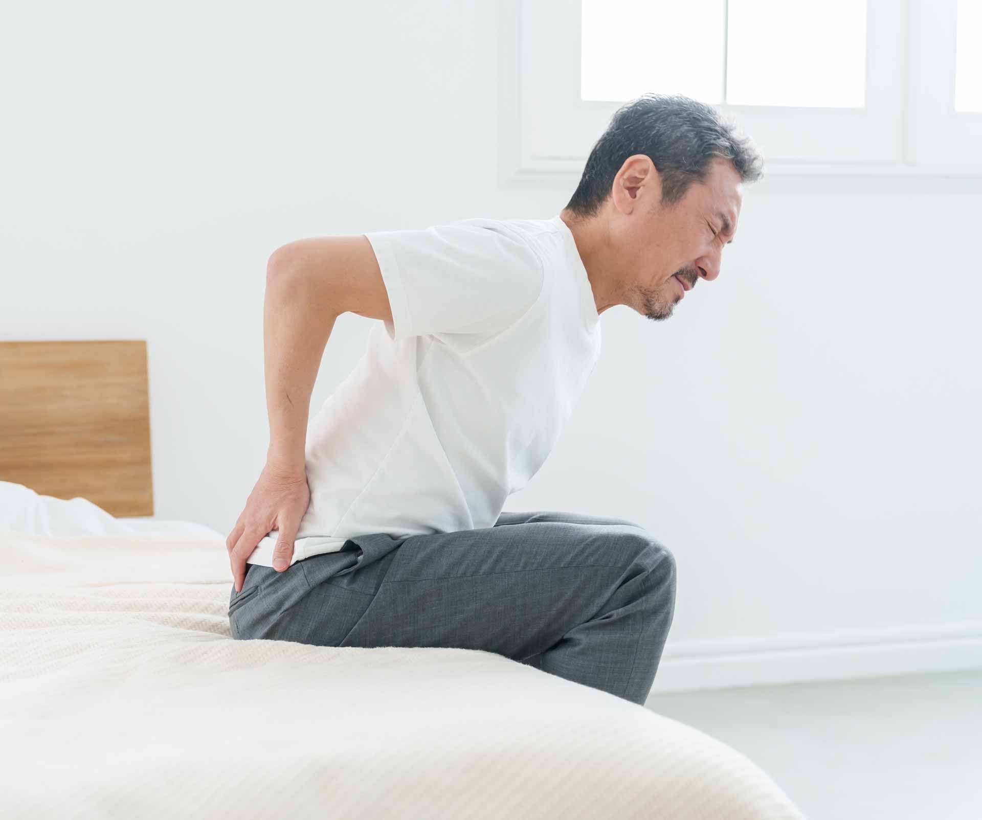 Man sitting on bed, holding his lower back in pain in a bright bedroom