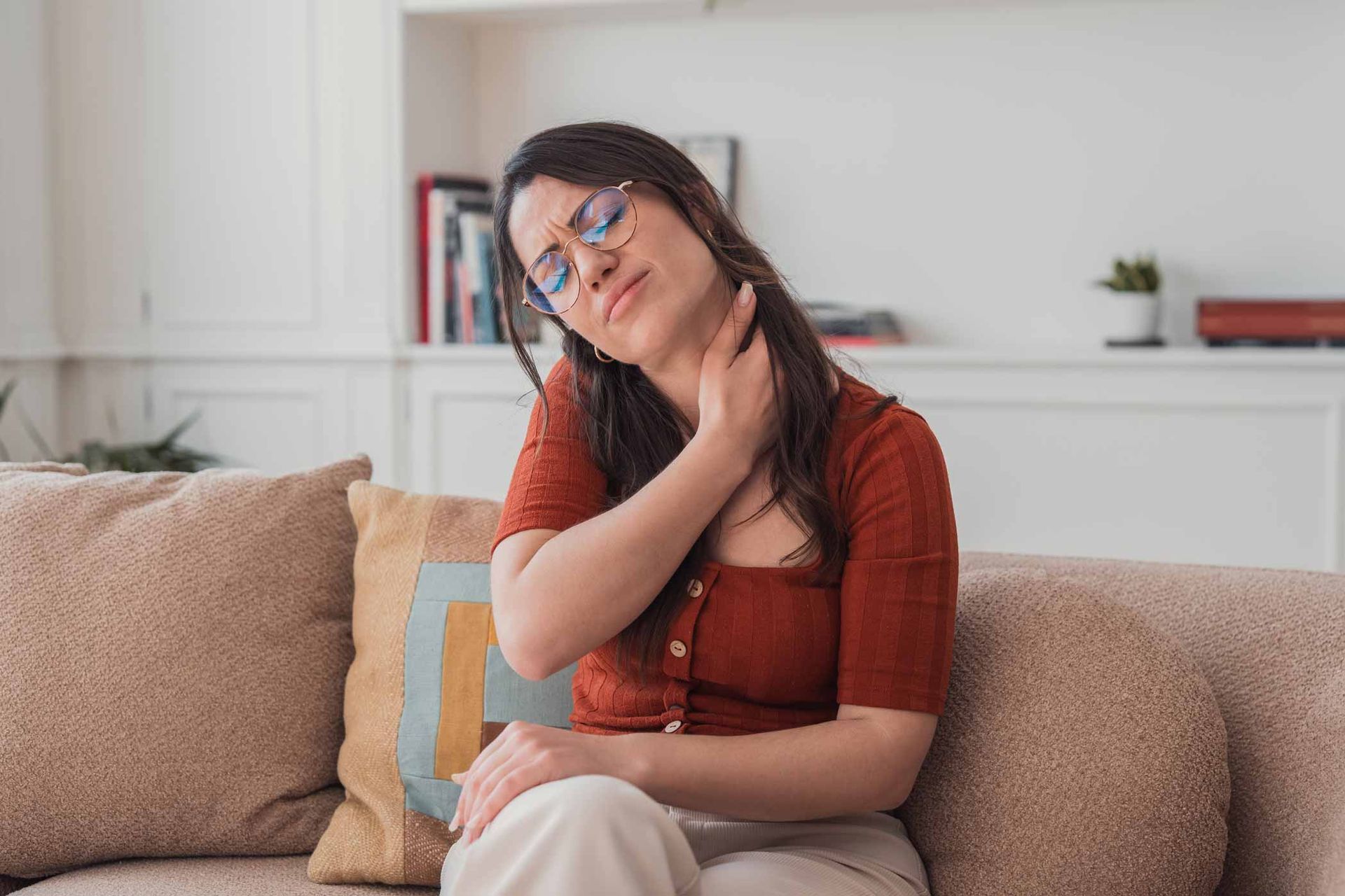 Person sitting on a beige couch, gently holding their neck in a bright living room.