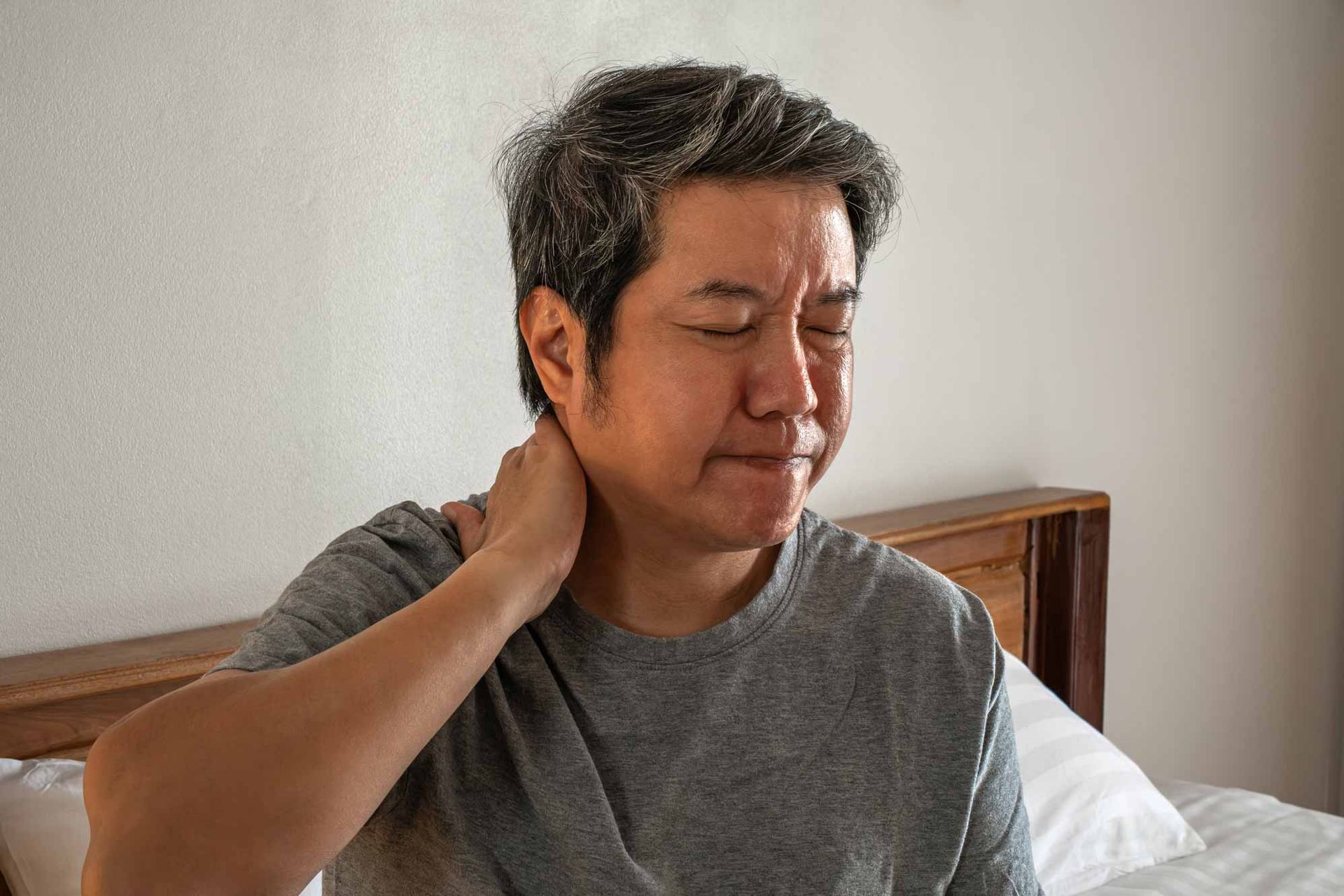 Person sitting on a bed, holding the back of their neck with eyes closed in discomfort.