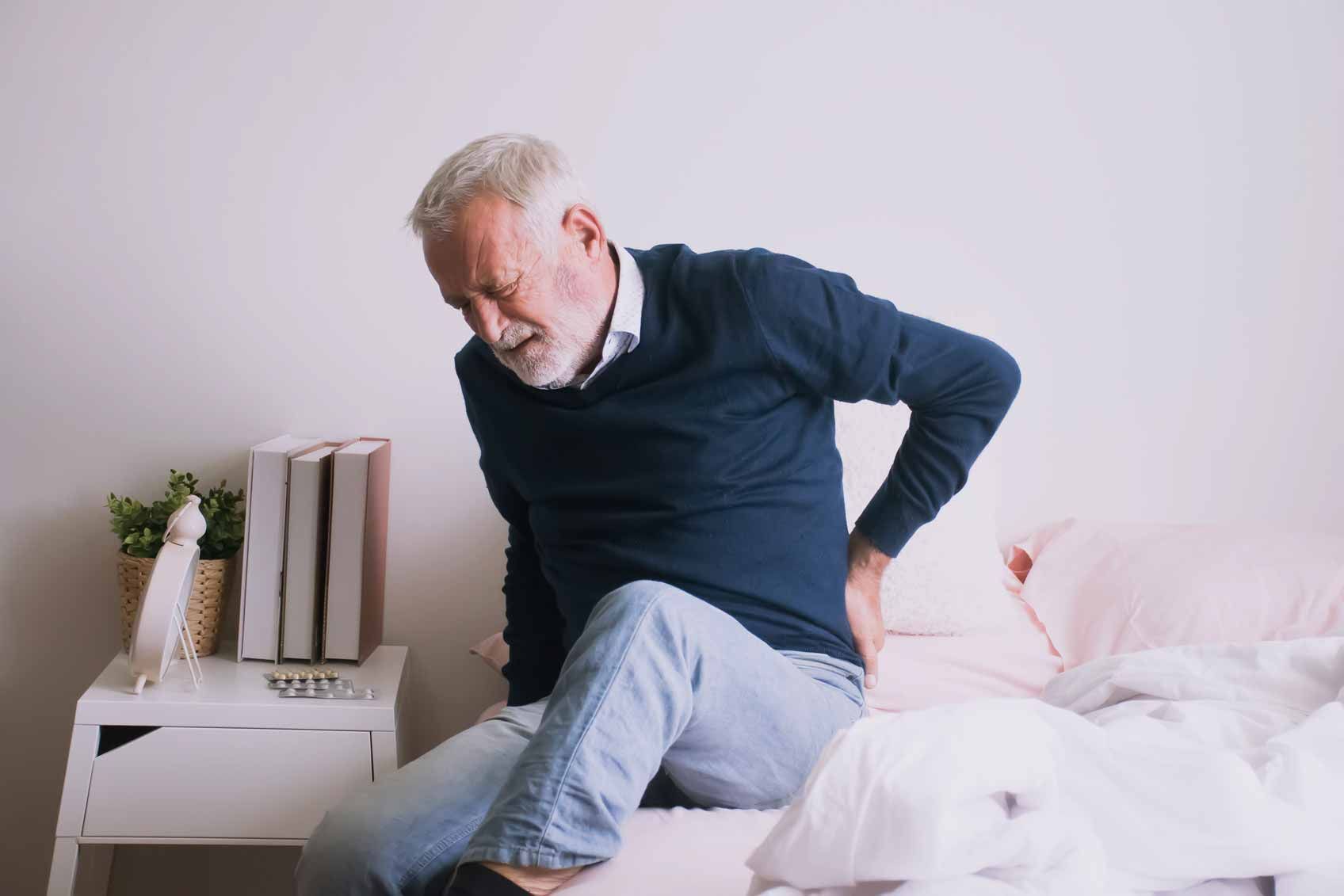 Person in a navy sweater sits on a bed, holding their lower back in apparent pain.