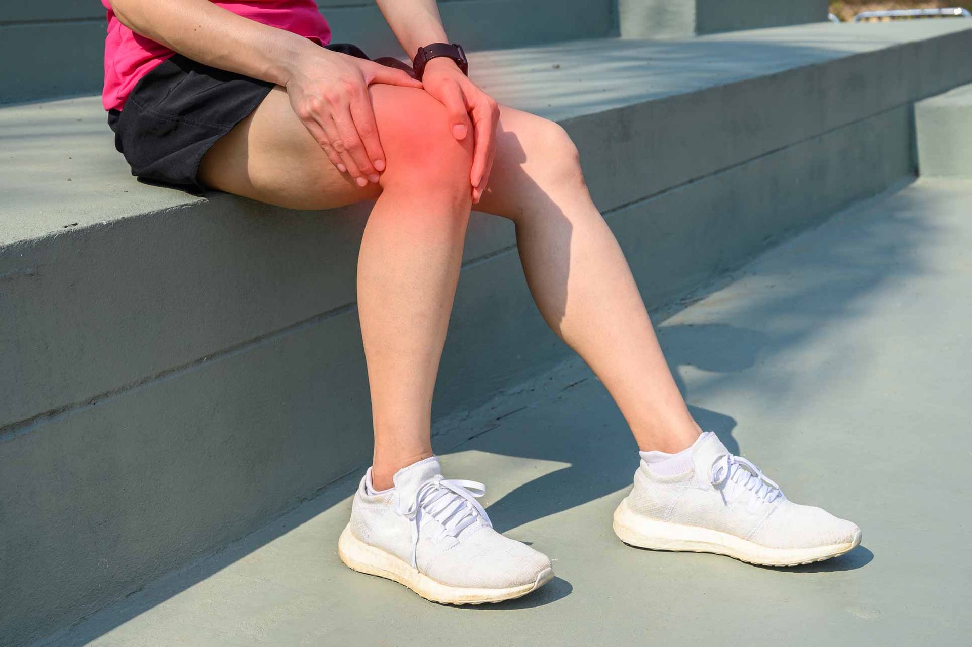 Person sitting on a concrete step, holding a red-highlighted knee, wearing black shorts and white sneakers.