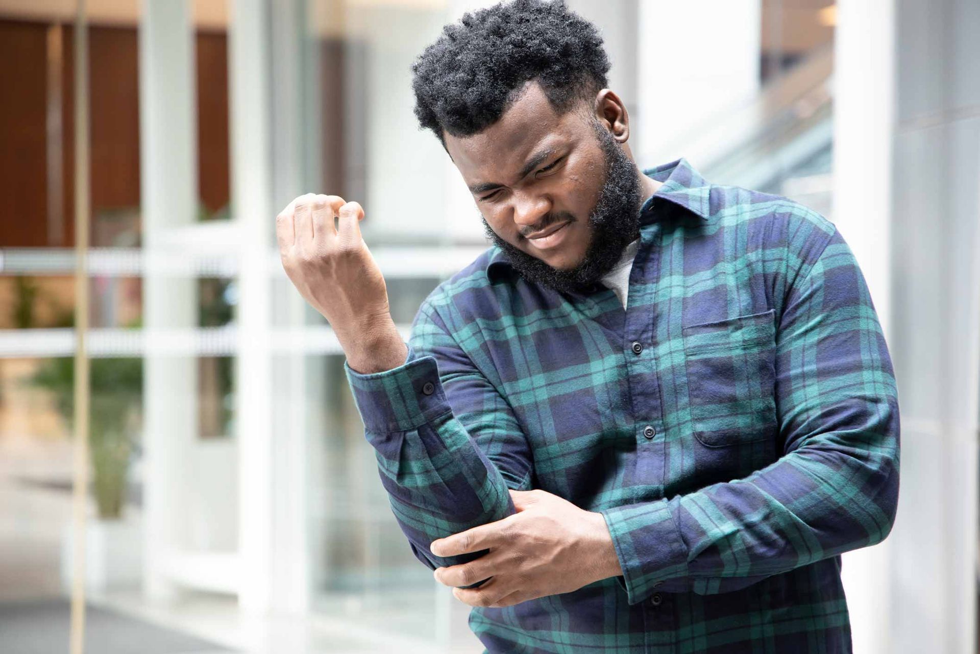 Man grimacing and clutching his elbow in pain, wearing a green plaid shirt outdoors.