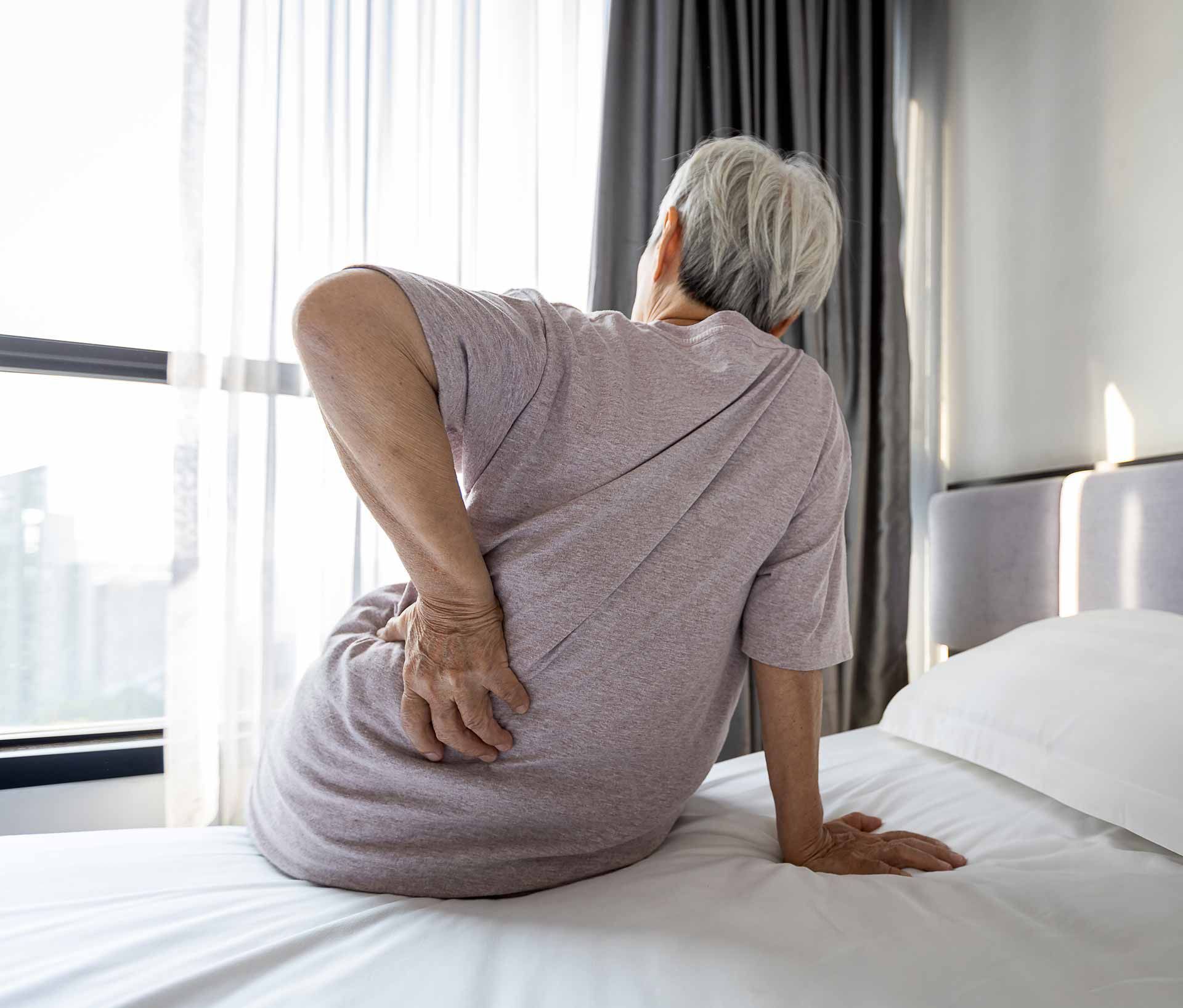 Person sitting on a bed, holding their lower back in a bright bedroom.