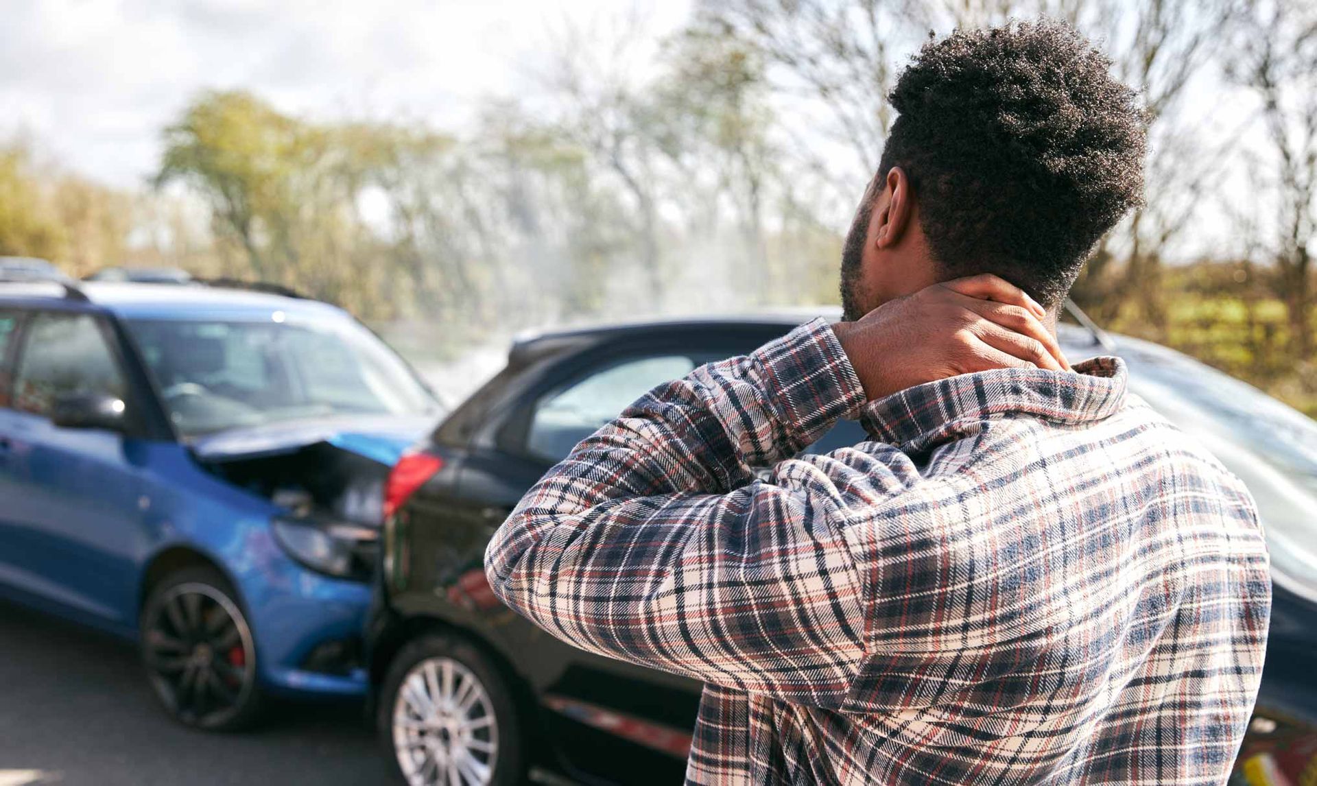 Two cars collide on a road while a person stands nearby holding their neck.