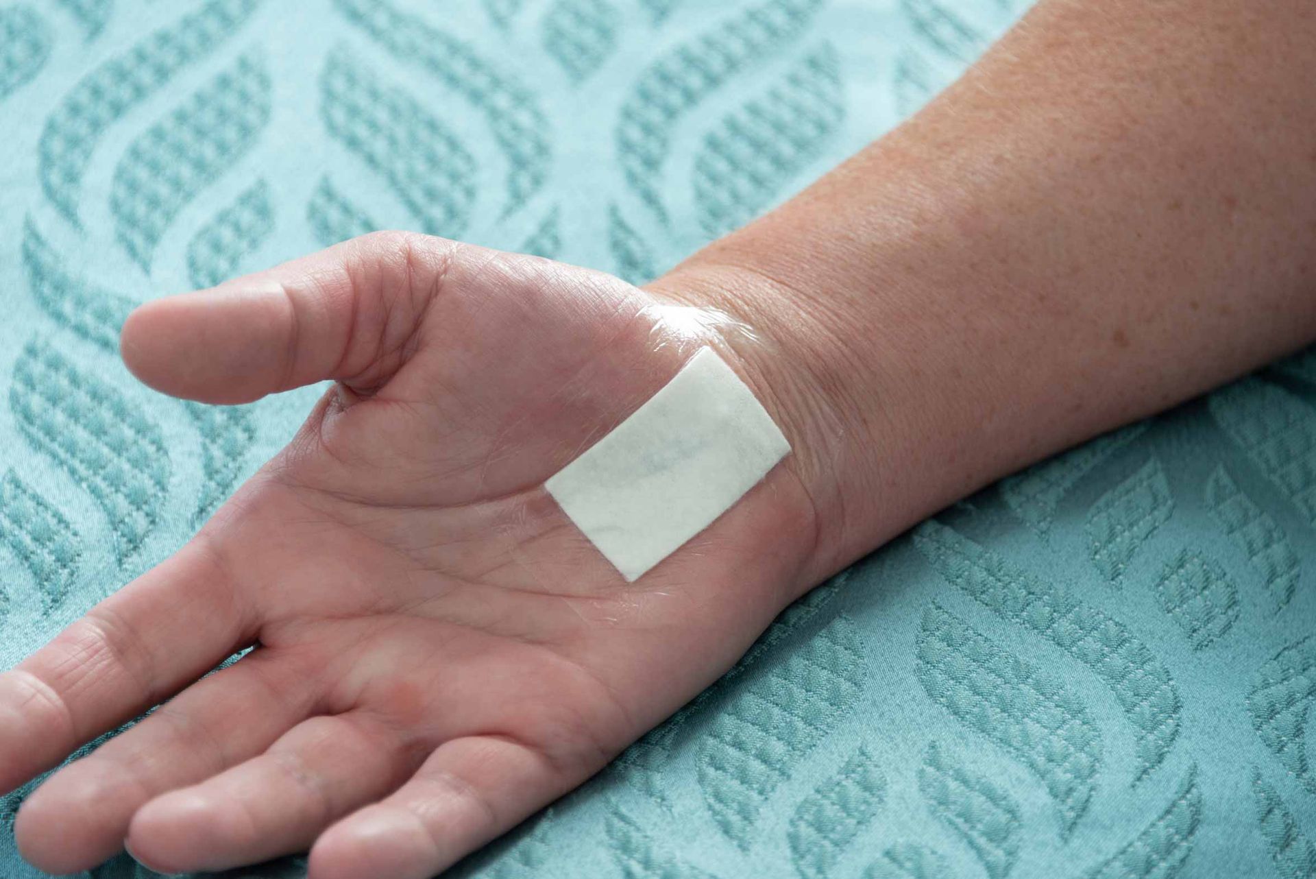 Hand with a small white bandage on the wrist, resting on a teal patterned towel.