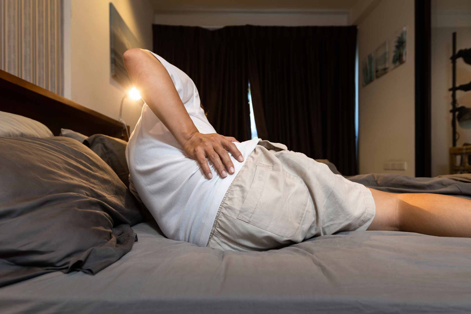 Person lying on a bed holding their lower back in a dimly lit bedroom