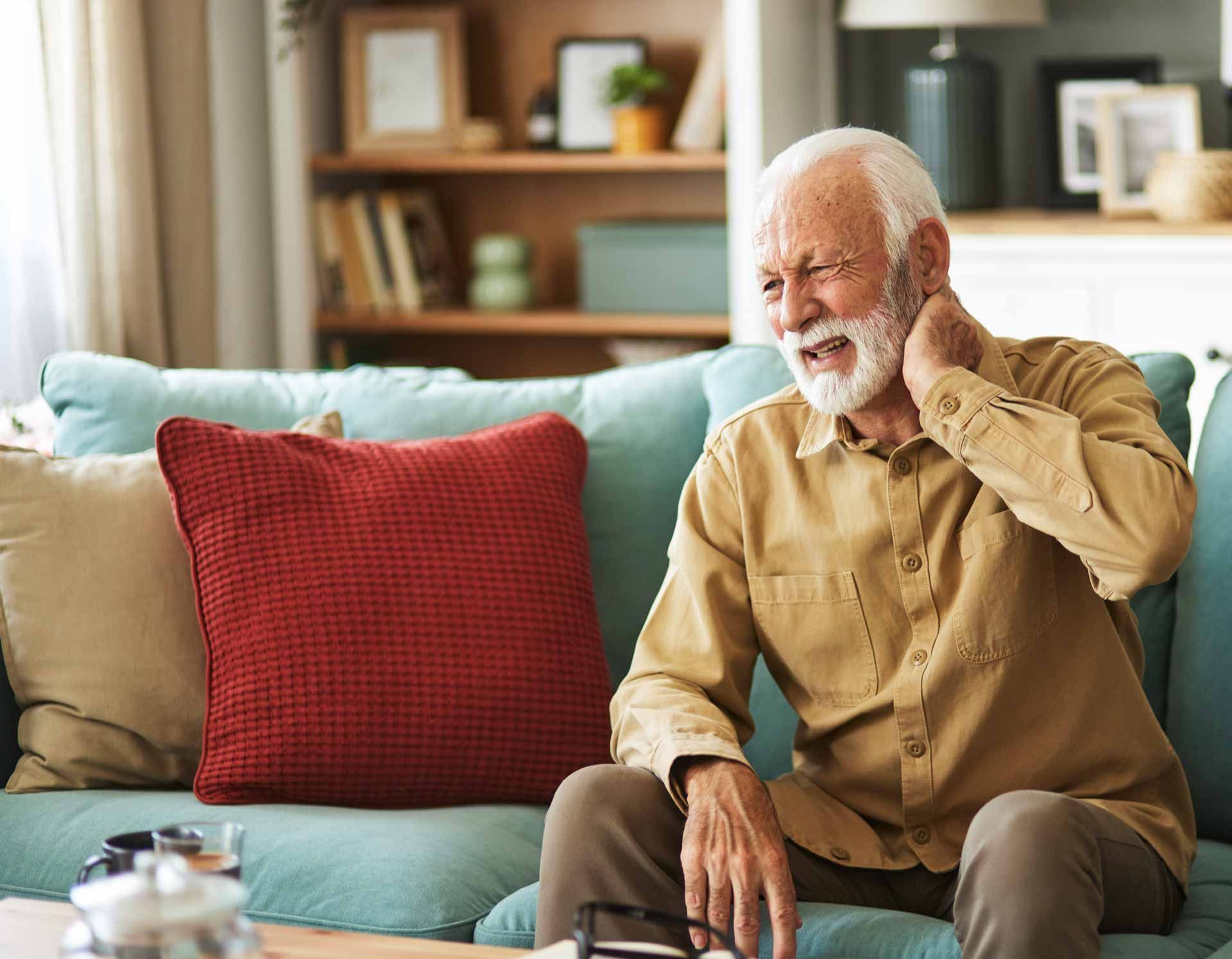 Elderly man sitting on a teal sofa, holding his neck in discomfort, with a red pillow beside him.