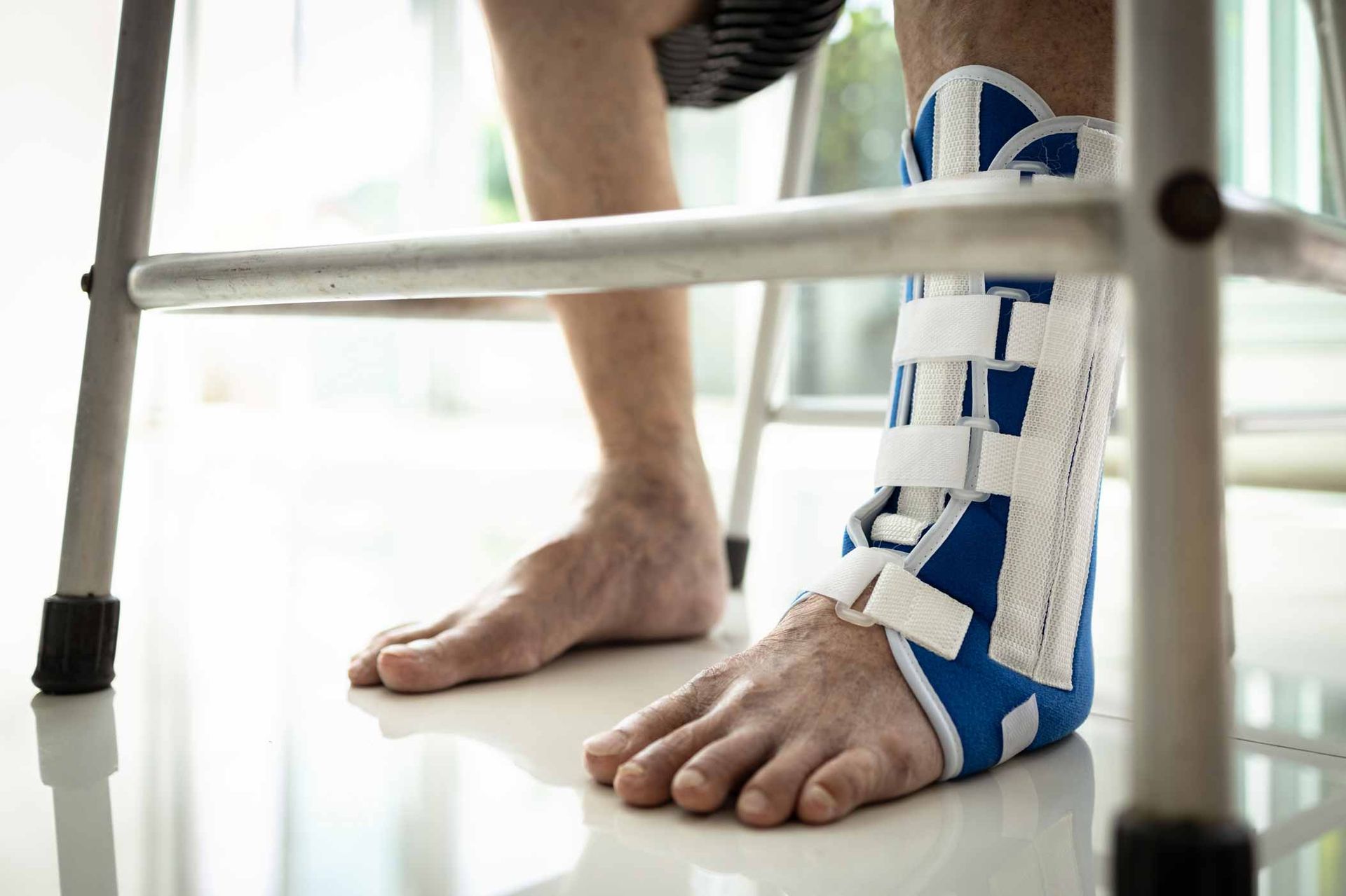 Person wearing a blue-and-white ankle brace beside a walker, standing on a bright floor.